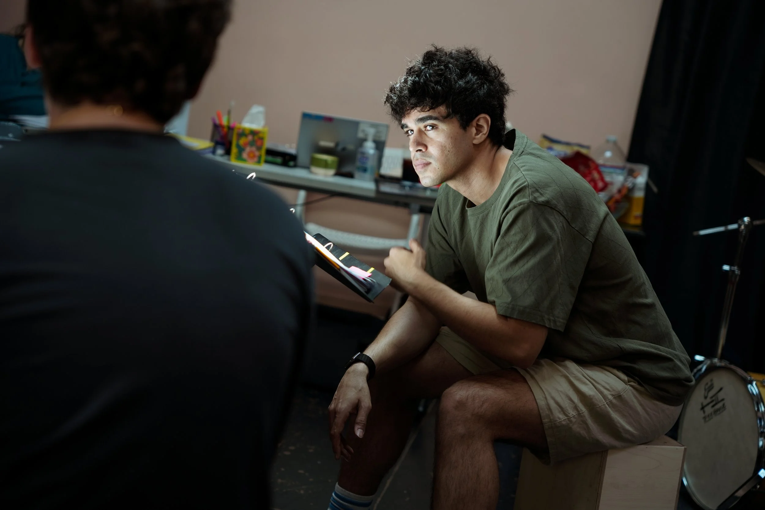 Young man with curly dark hair sitting on a drum in a room, looking intensely at someone out of view, with a table and various objects in the background.