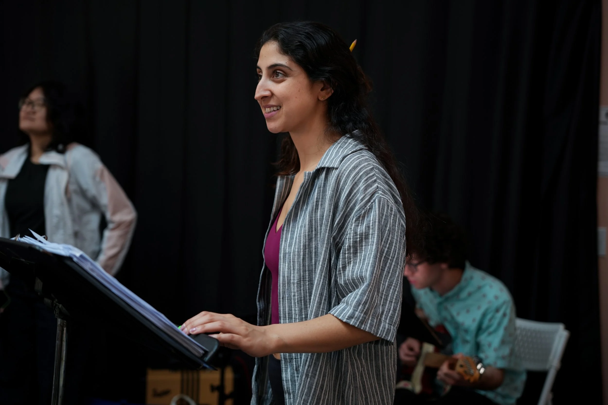 A woman with dark hair smiling while playing a keyboard. She is wearing a gray striped blazer and a purple top. In the background, another woman with glasses and a patterned shirt and a man with glasses and a patterned shirt are also present.