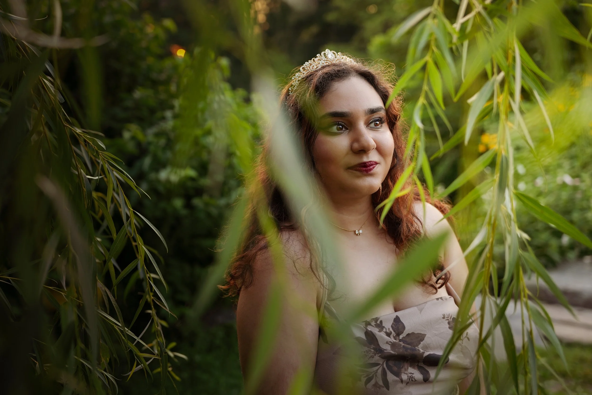 A woman with brown hair and a tiara, standing outdoors among green foliage, wearing a strapless dress with floral pattern details.