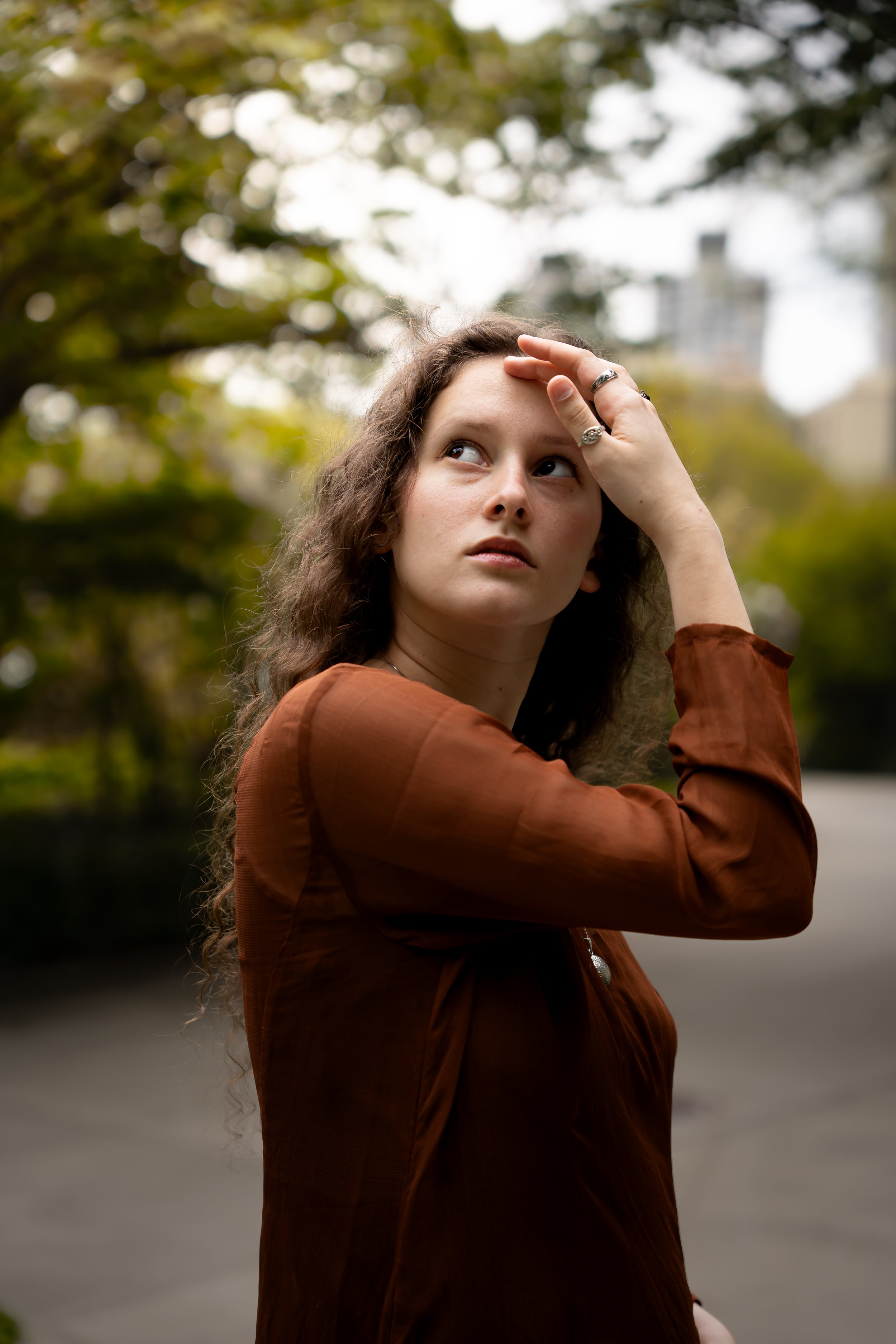 A young woman with long, curly brown hair, wearing a brown jacket and rings on her fingers, is outdoors with blurred trees and buildings in the background. She is touching her forehead with her right hand and looking to the side.