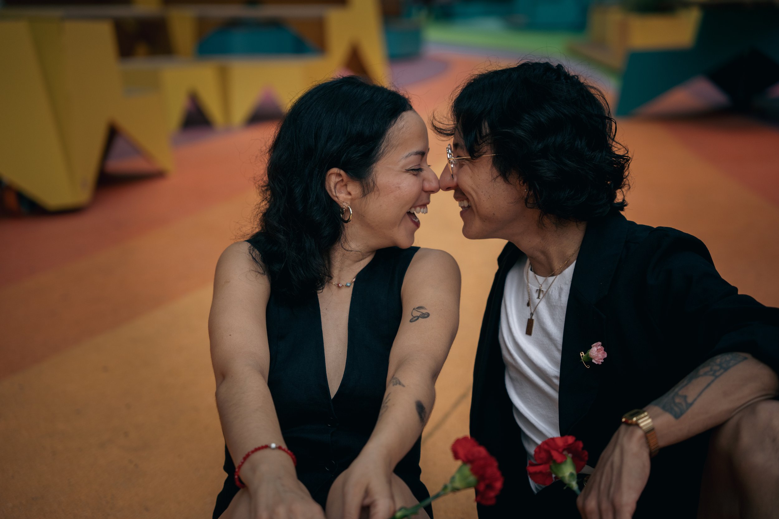 A couple with their foreheads touching, smiling, and holding red carnations.