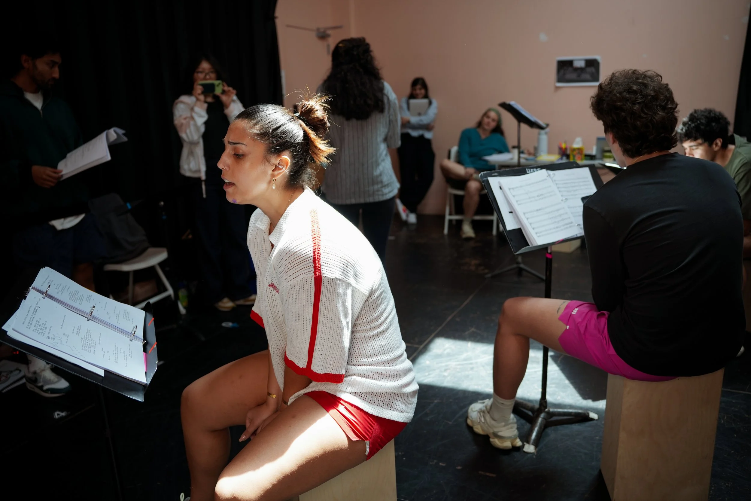 A woman dressed in a white and red athletic shirt and red shorts, sitting on a wooden stool, singing or speaking with her eyes closed, during a rehearsal or singing session. Several people are visible in the background, some sitting and some standing