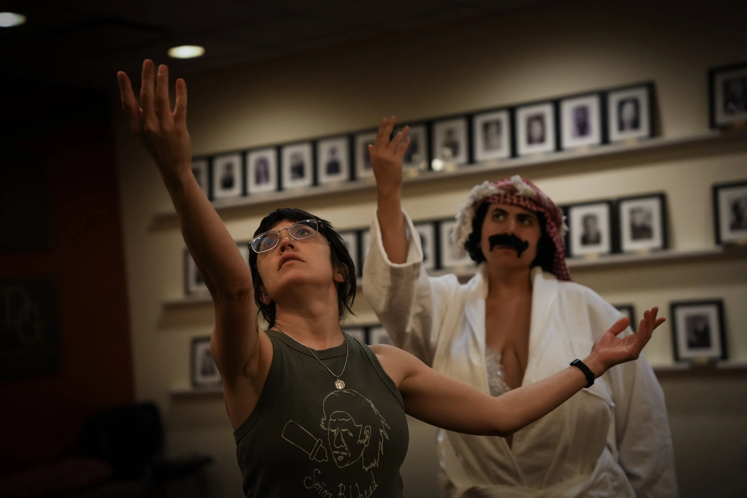 Two individuals dance together in a room decorated with framed photographs on the wall, one wearing glasses and a sleeveless shirt with a drawing of a man's face, the other dressed in a white costume with a red and white headscarf and a fake mustache
