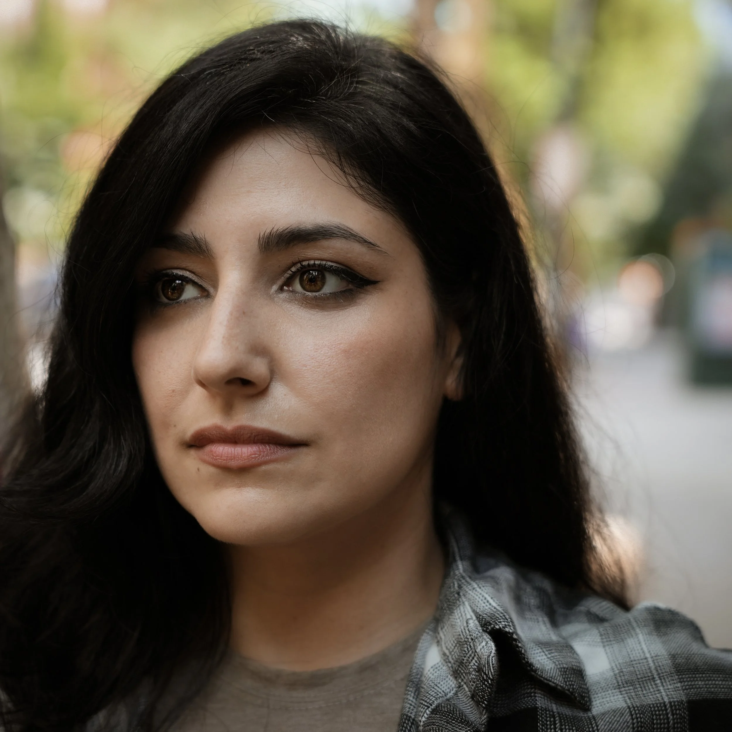 Partial view of a young woman with long dark hair and brown eyes, wearing a checkered shirt, outdoors with blurred greenery background.