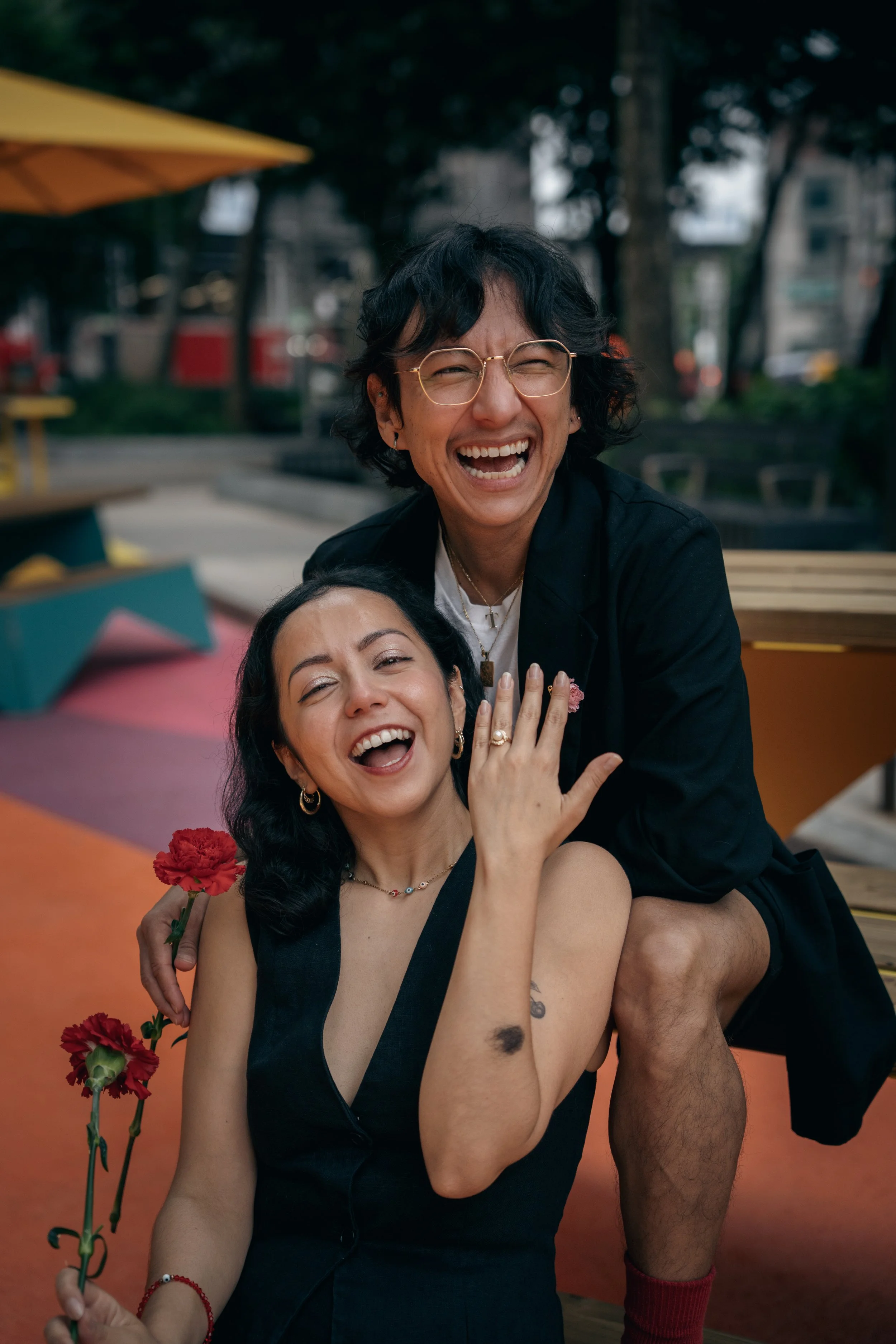 Two women smiling and posing for a photo outdoors, with one holding red carnations, in a casual setting with trees and a cityscape in the background.
