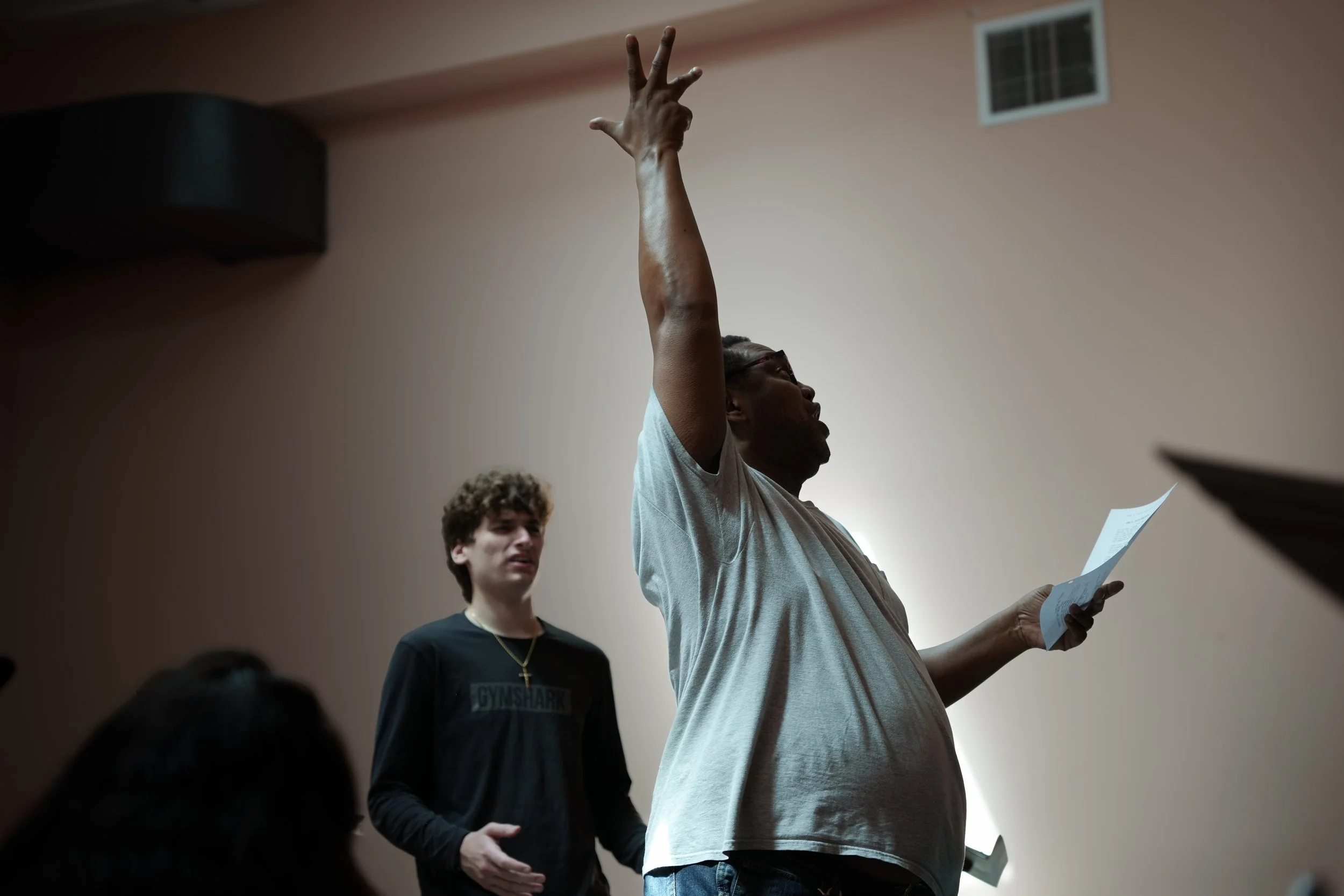 A person with glasses raising their hand with two fingers in a peace sign during a meeting or classroom setting, with another person standing nearby holding a paper, in a room with a beige wall and a window.