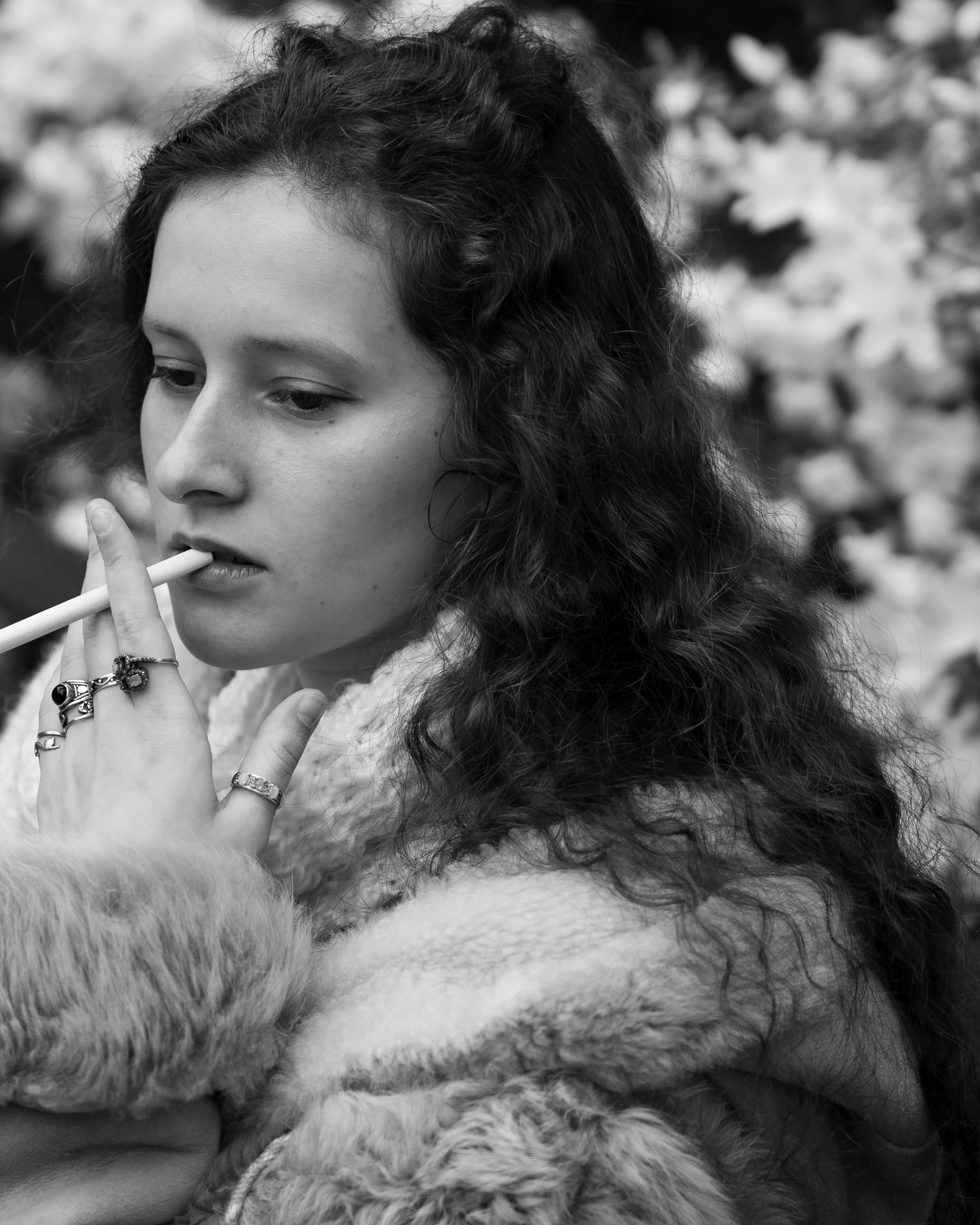 A black-and-white close-up photo of a woman with curly hair holding a cigarette near her lips, wearing multiple rings on her fingers, with a blurred floral background and a fluffy fur garment or blanket around her.