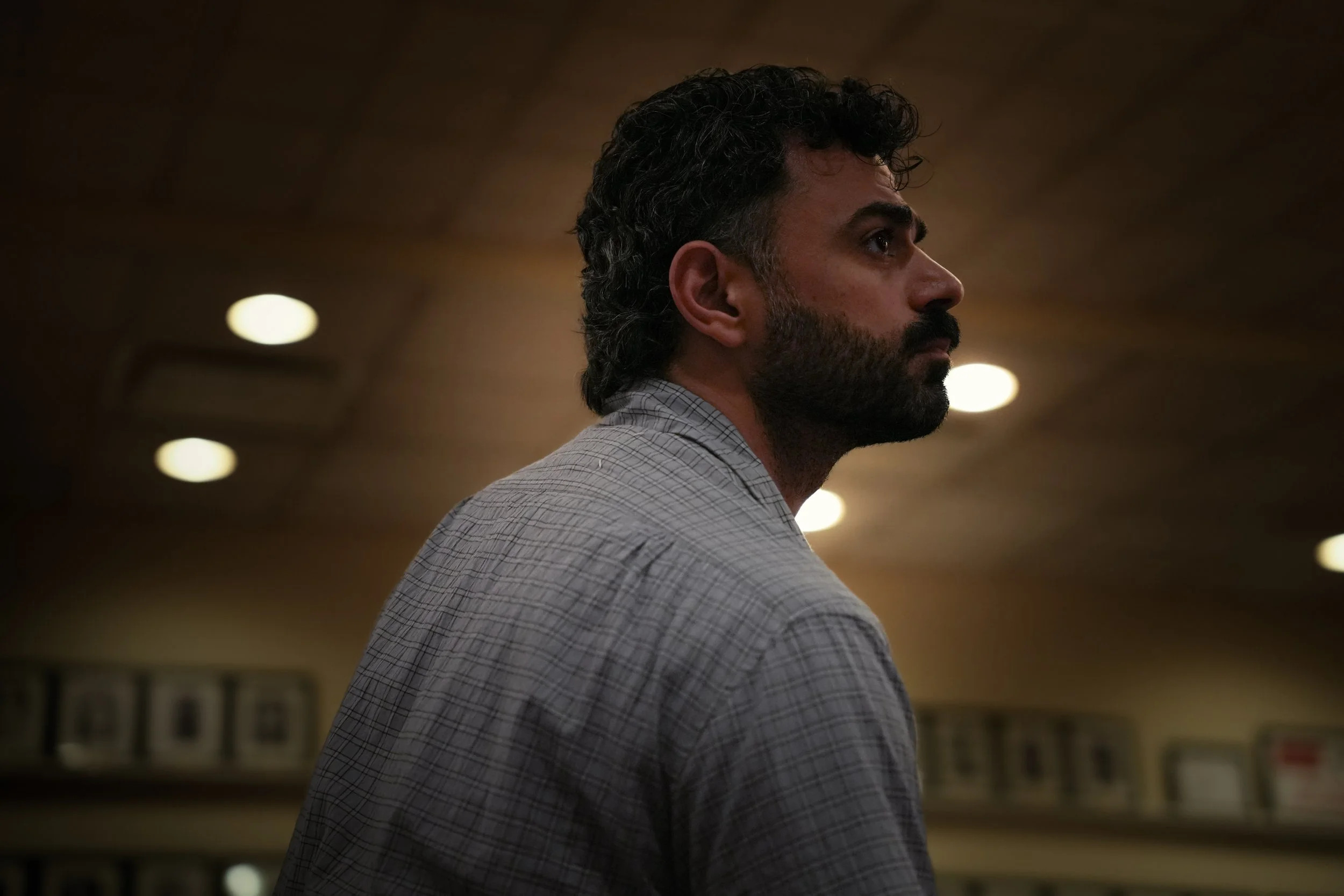 Profile side view of a bearded man with dark, curly hair, wearing a checkered shirt, indoors with blurred lights and background.