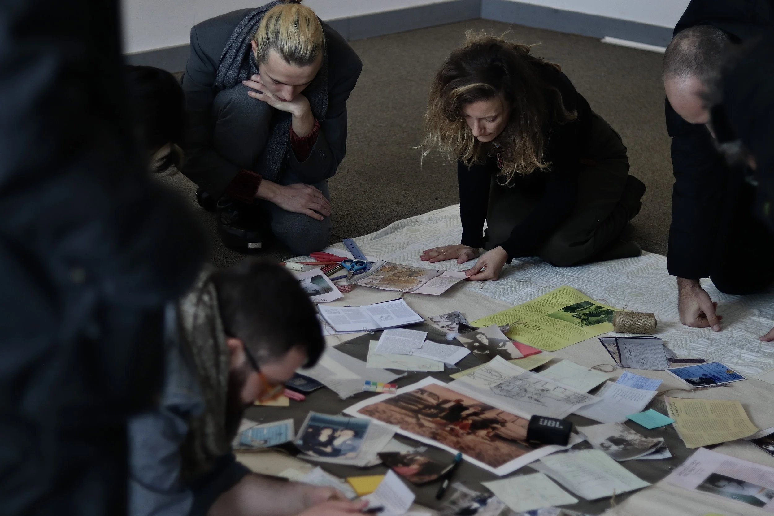 People kneeling on the floor, organizing papers and books, with a variety of documents, magazines, and office supplies spread out on the surface.