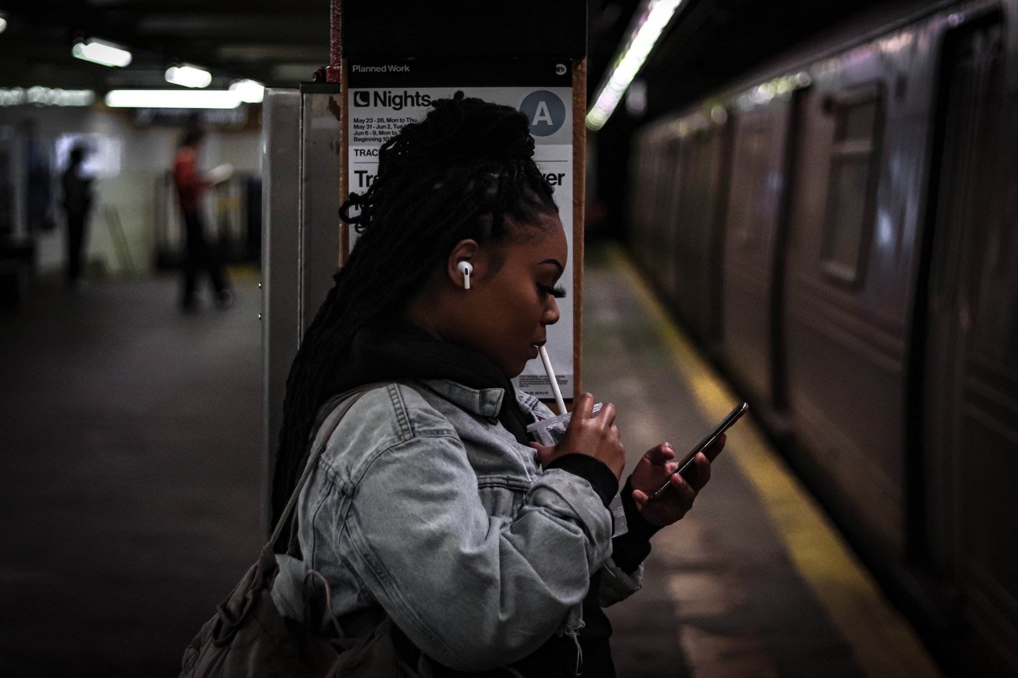 Person with earbuds standing in a subway station, drinking from a cup and looking at a phone. Bronx harlem nyc