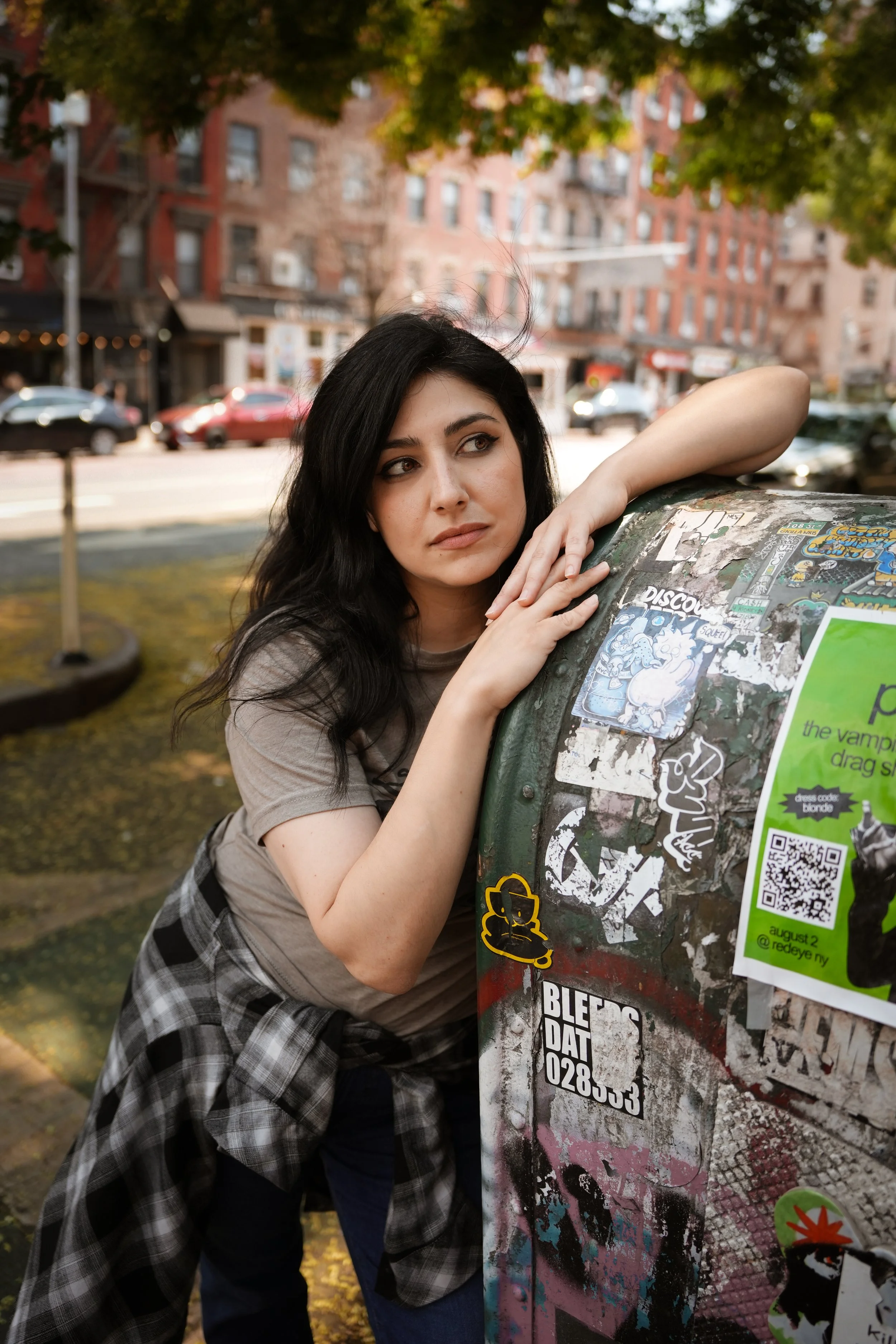 A woman with long black hair leaning on a decorated, sticker-covered street mailbox in an urban setting with brick buildings and parked cars in the background.