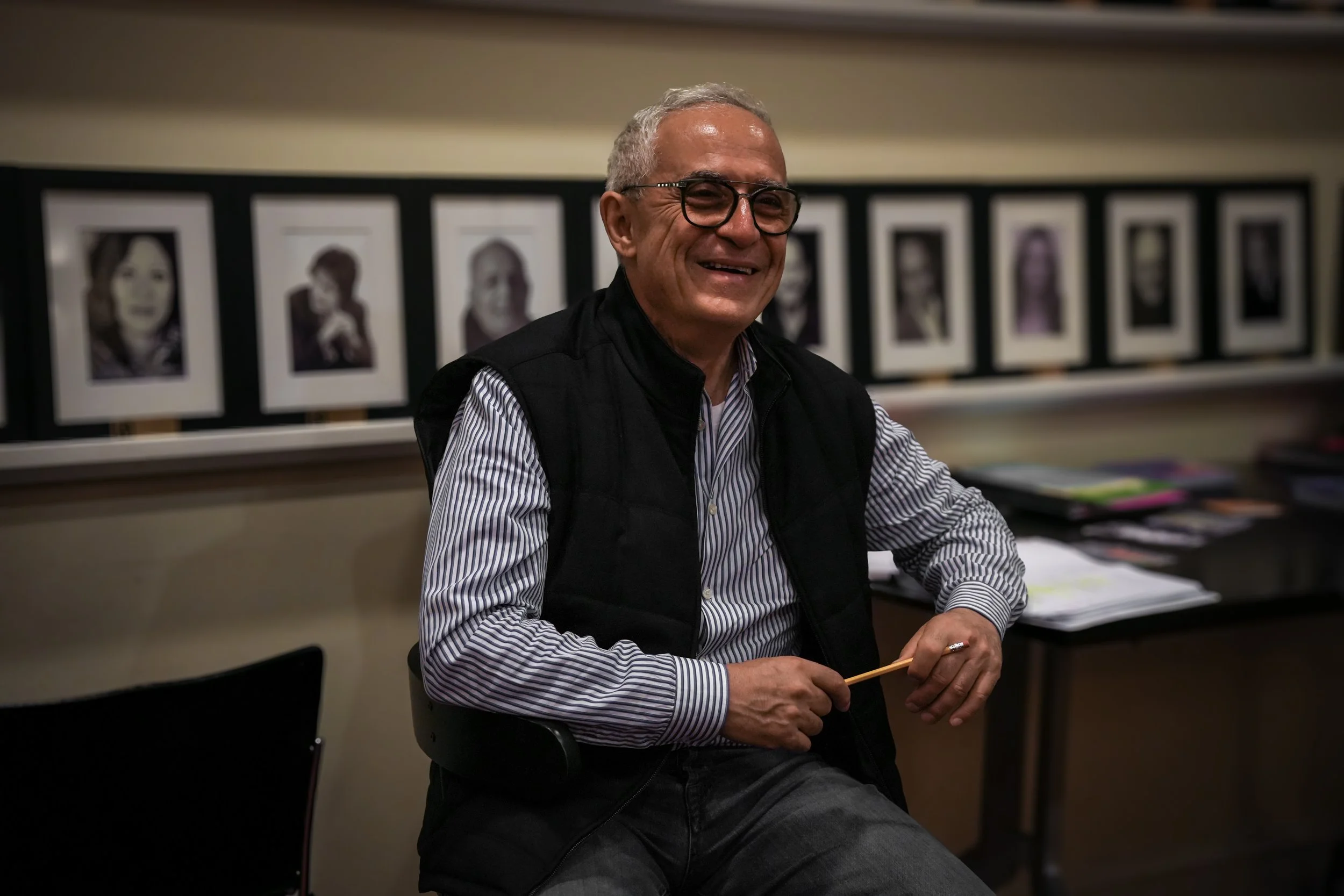 An older man with gray hair and glasses, smiling, sitting at a desk holding a pencil in his right hand, with black and white portraits on the wall behind him.