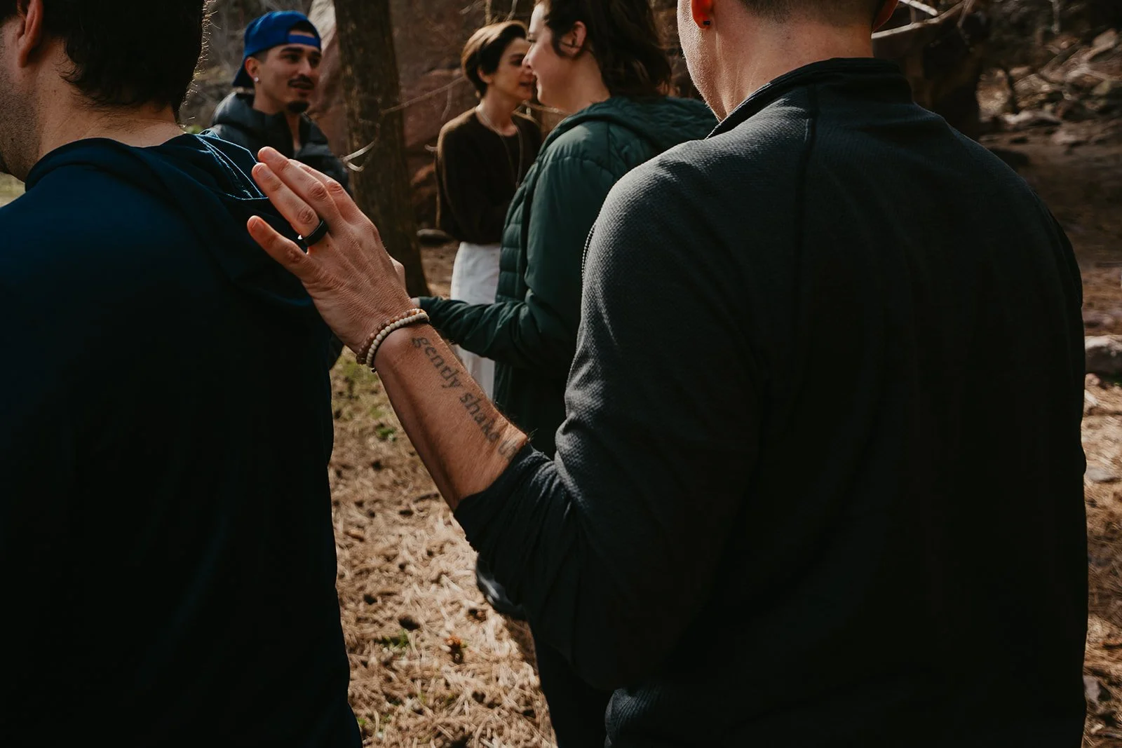 A group of people outdoors in a wooded area, engaged in conversation. Focus on a man touching a woman's shoulder, with others in the background.