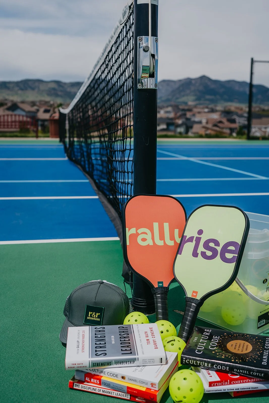 Collection of sports equipment and books on a tennis court, including two paddles, tennis balls, a cap, and books about leadership, culture, and markets.