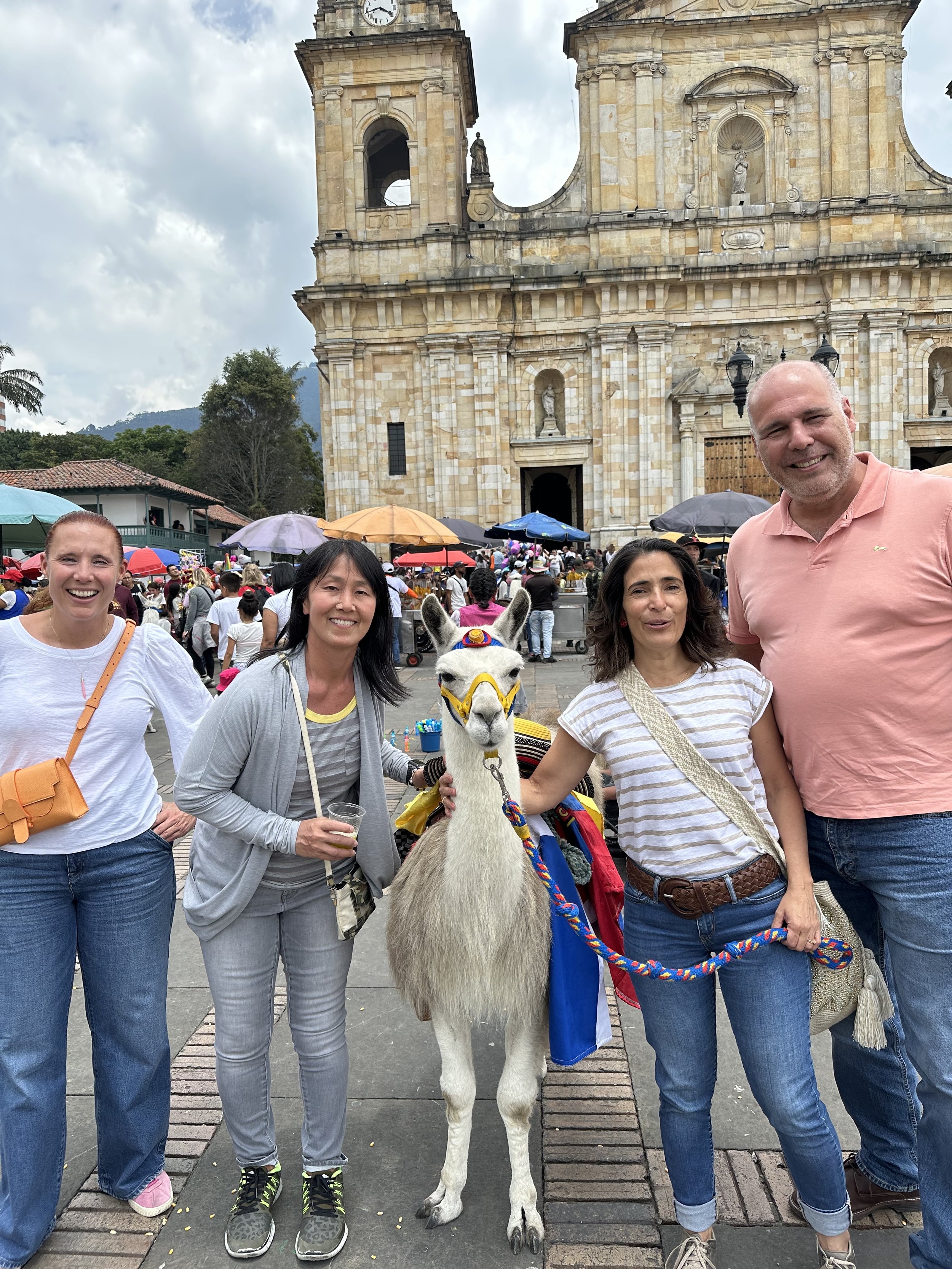 Four people standing in front of a historical building with a llama dressed in colorful accessories, at a crowded outdoor event.