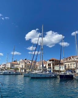 A marina with sailboats docked on calm water, sailboats with masts, and residential buildings under a bright blue sky with white clouds.