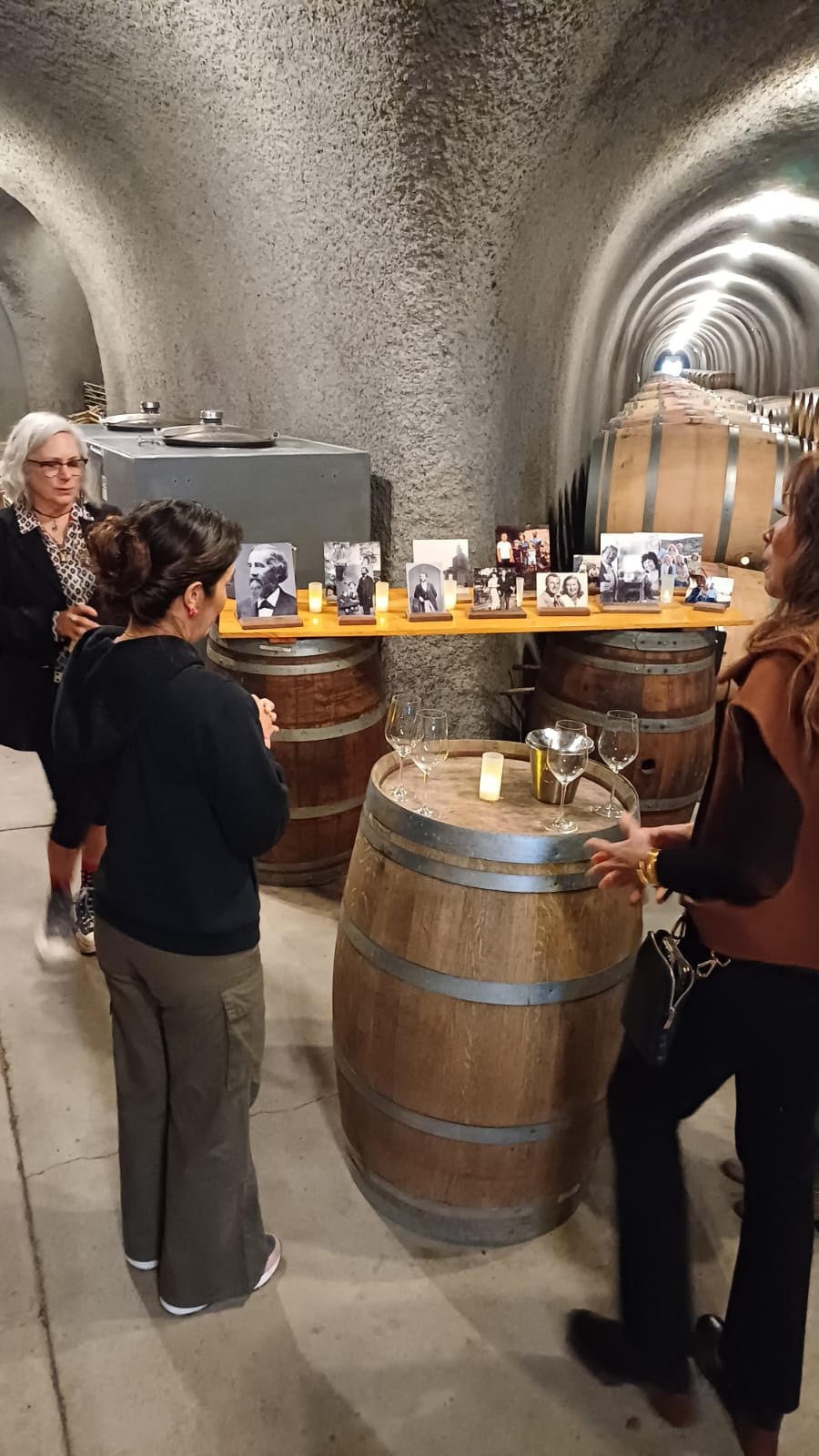 Three women gather around a table made from a wine barrel in a wine cellar with large barrels and a framed display of black-and-white photographs behind them.