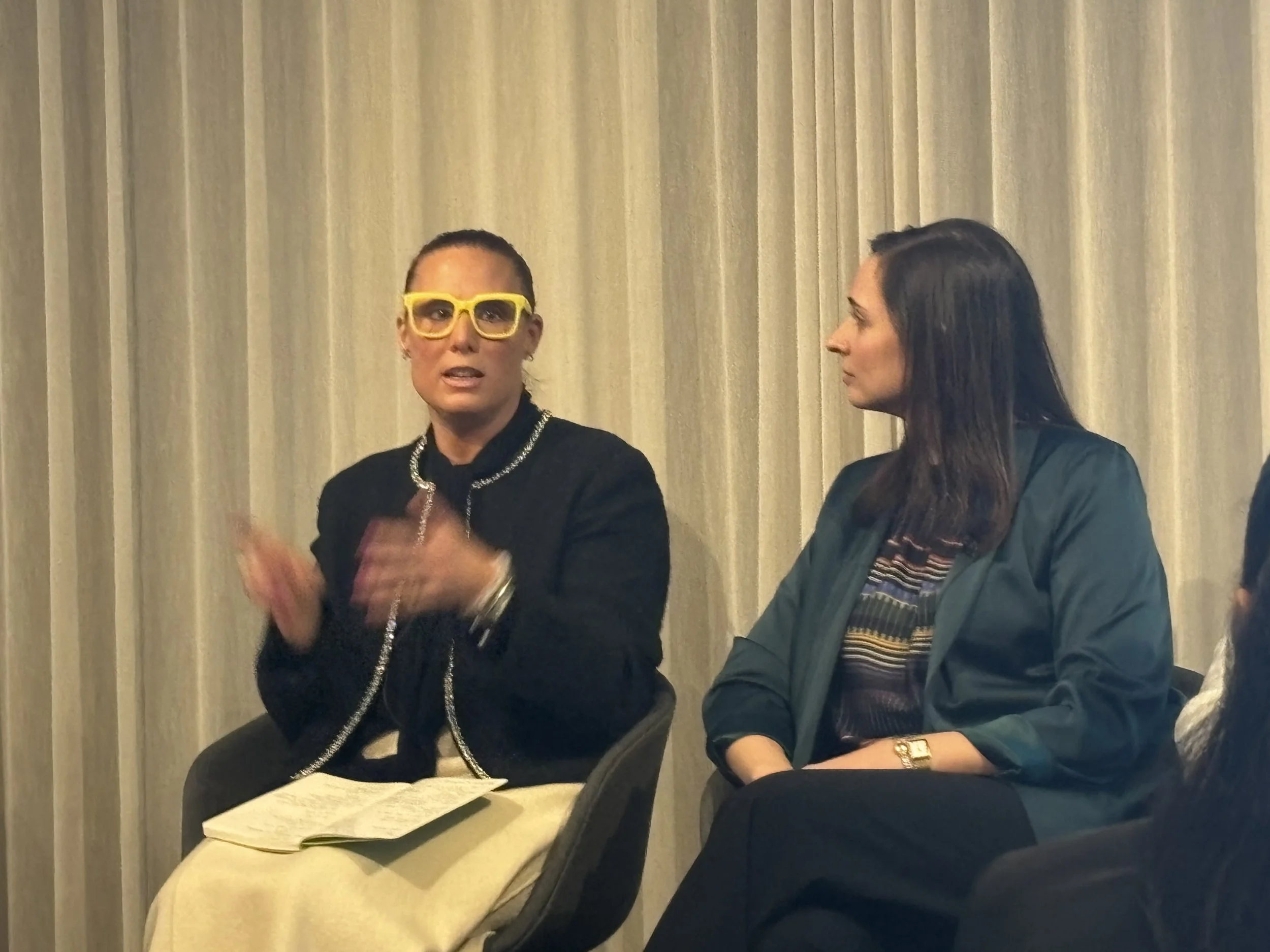 Two women sitting and engaged in conversation, with one woman wearing yellow glasses and a black jacket, and the other woman wearing a green jacket and patterned shirt, against beige curtains.