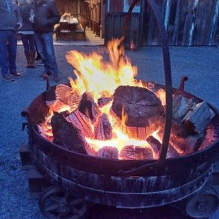 A large outdoor fire pit with burning logs and flames, surrounded by an urban setting.