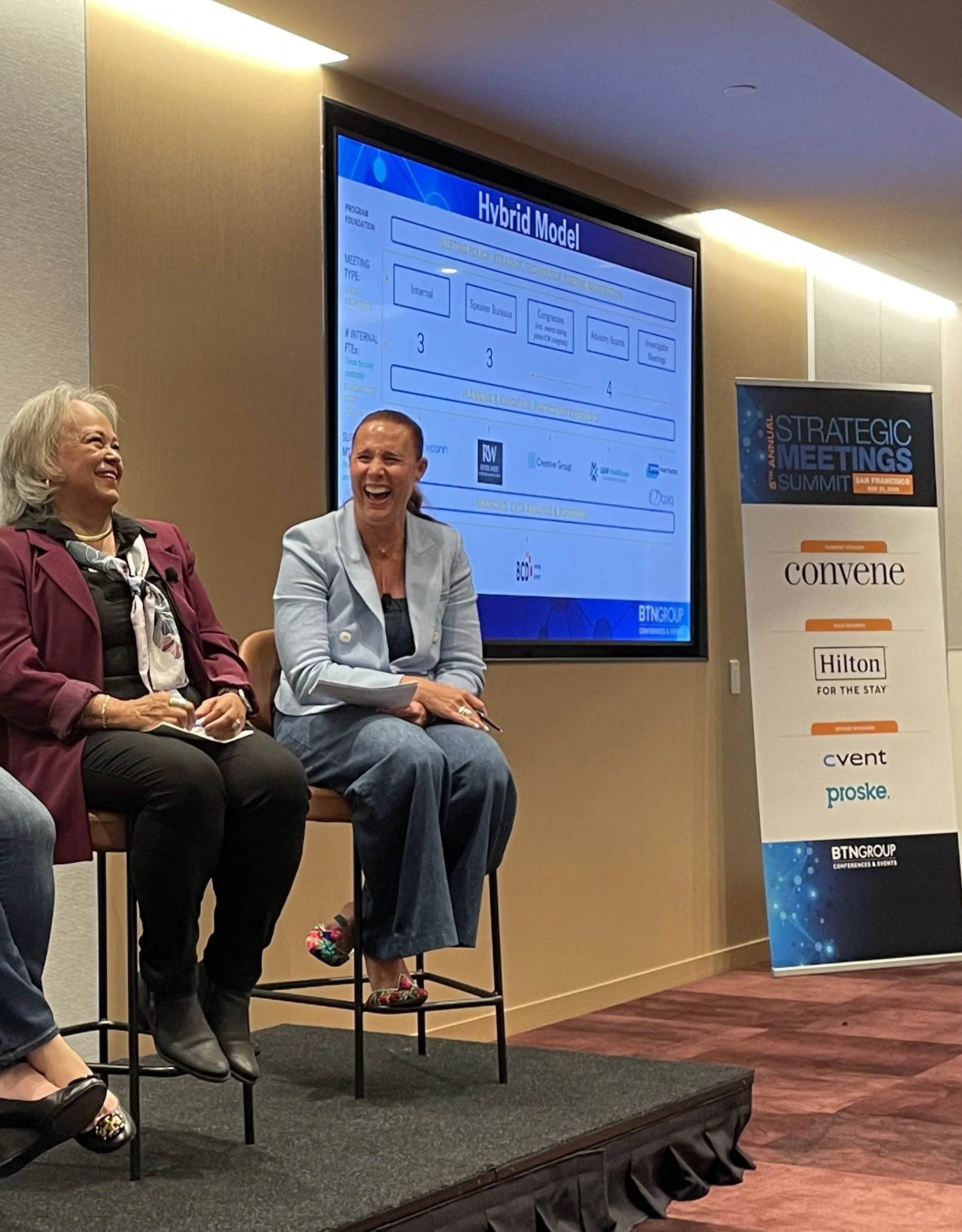 Two women sitting on stage at a conference, laughing. Behind them is a large screen displaying a slide titled 'Hybrid Model.' To the right is a vertical banner with conference information and sponsor logos.
