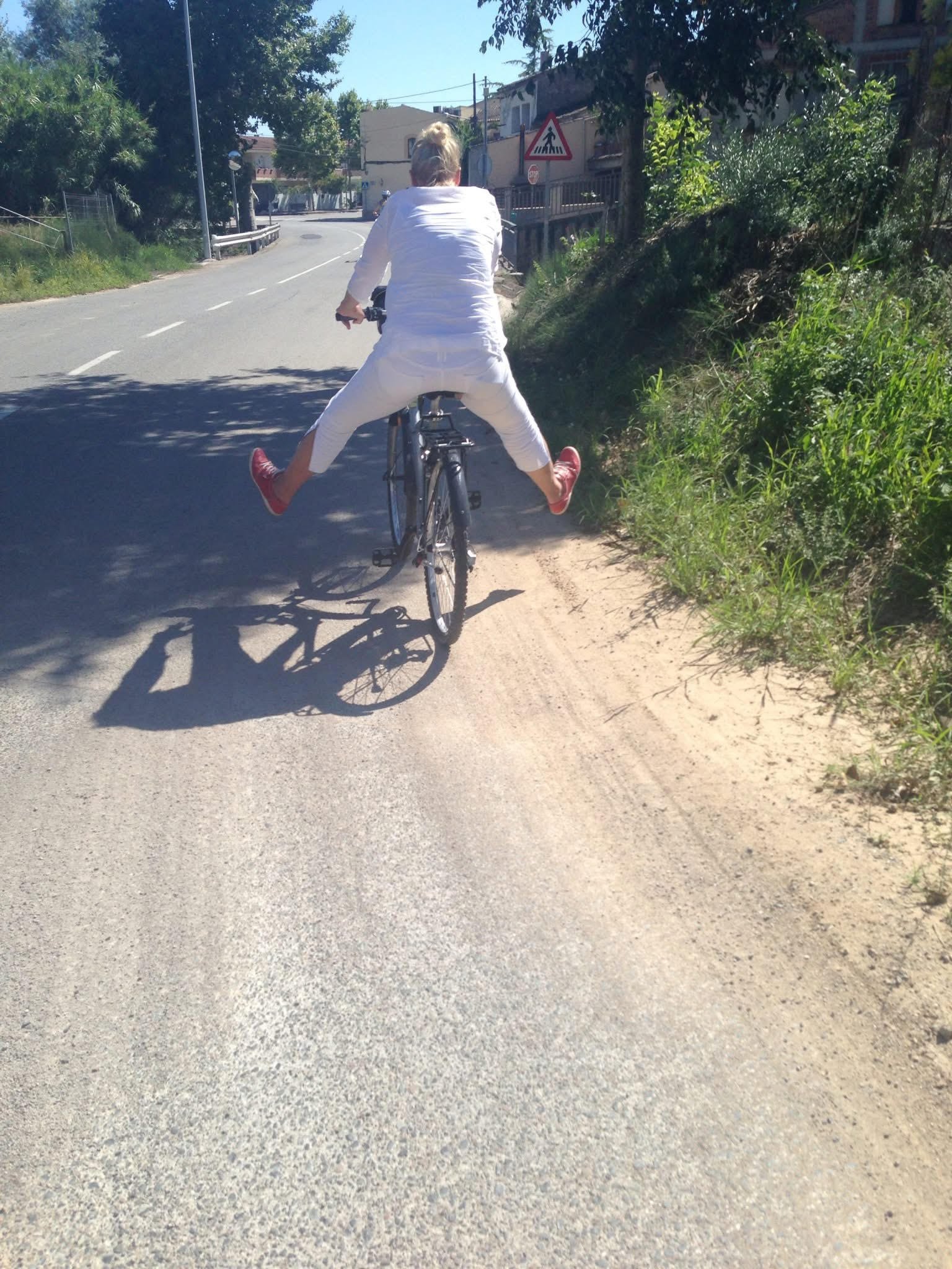A person riding a bicycle on a paved road with their legs spread wide apart, wearing white clothing and red shoes, casting a shadow on the road. The scene includes greenery on the side of the road and a traffic sign in the background.