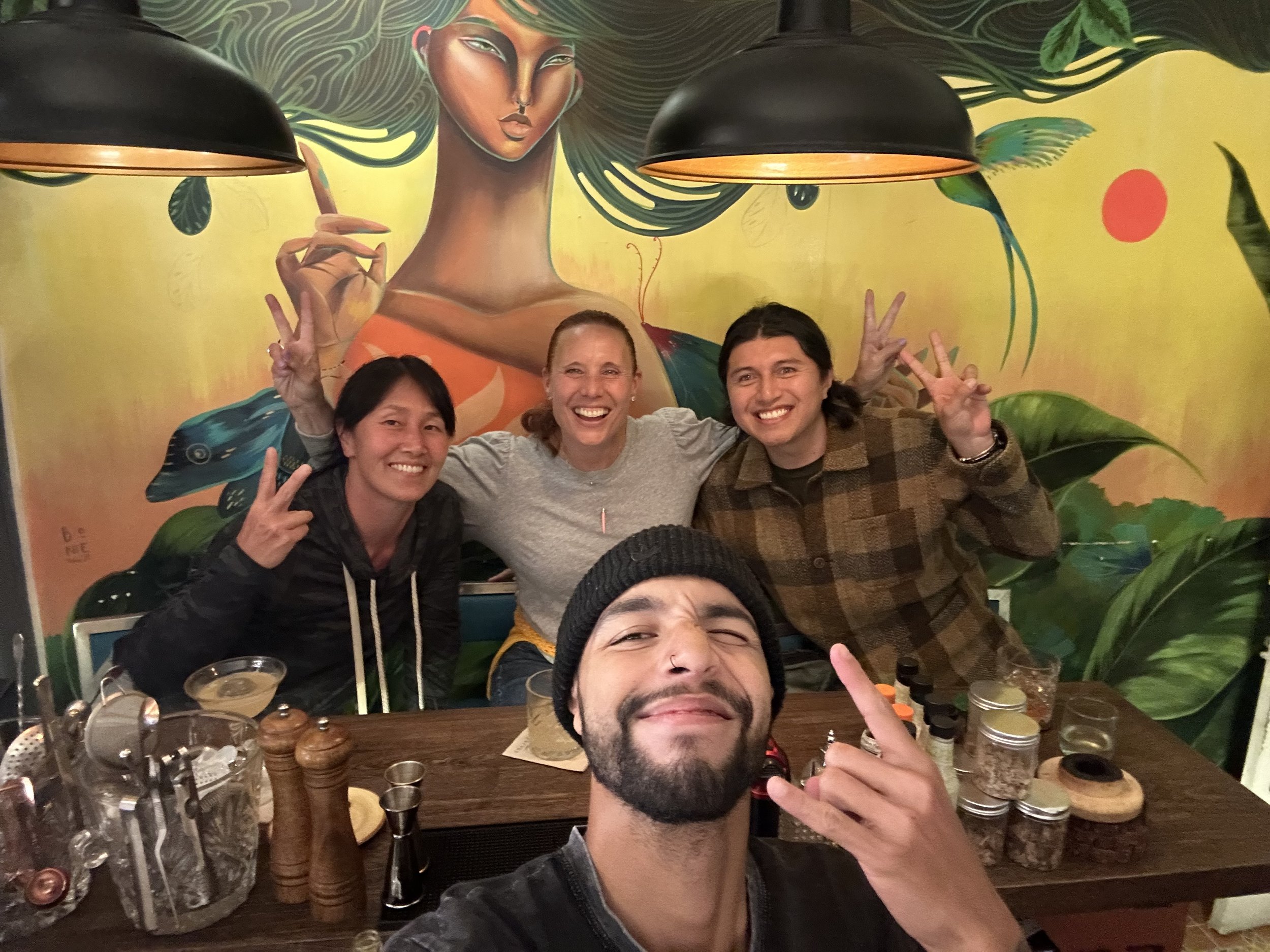 Four friends smiling and making peace signs at a table in a restaurant with a colorful mural of a woman with long hair in the background.