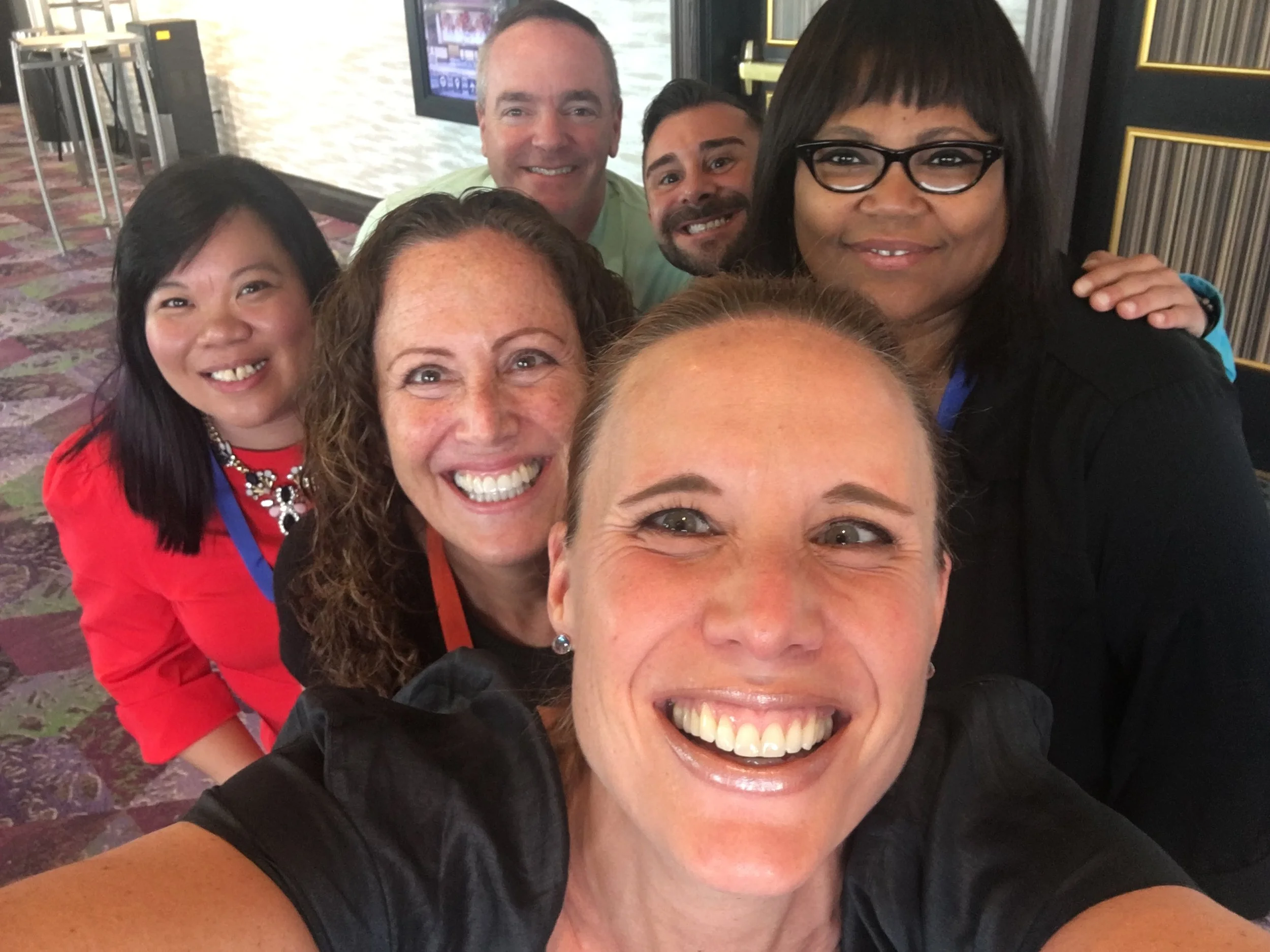 Group of seven smiling people taking a selfie at a social event or conference, with a colorful carpeted floor in the background.