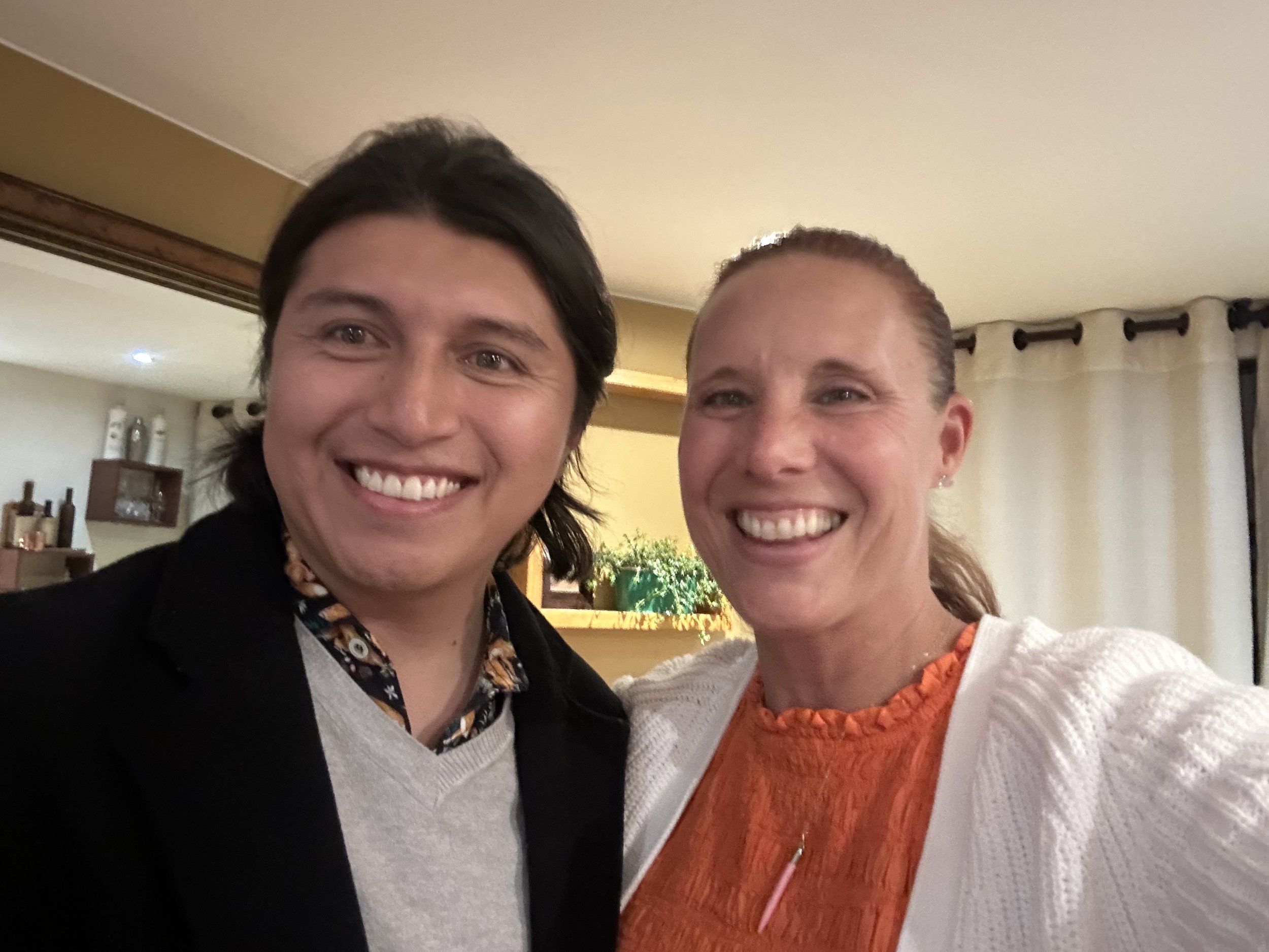 A man and woman smiling for a selfie indoors, with a background of curtains and a shelf with bottles.