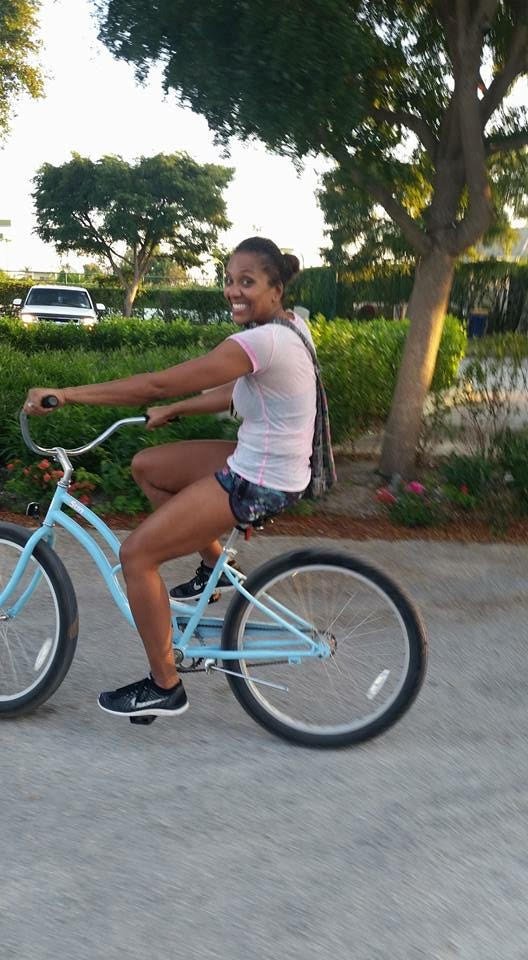 A woman is riding a light blue bicycle along a suburban street during daylight, smiling, with trees and greenery in the background.
