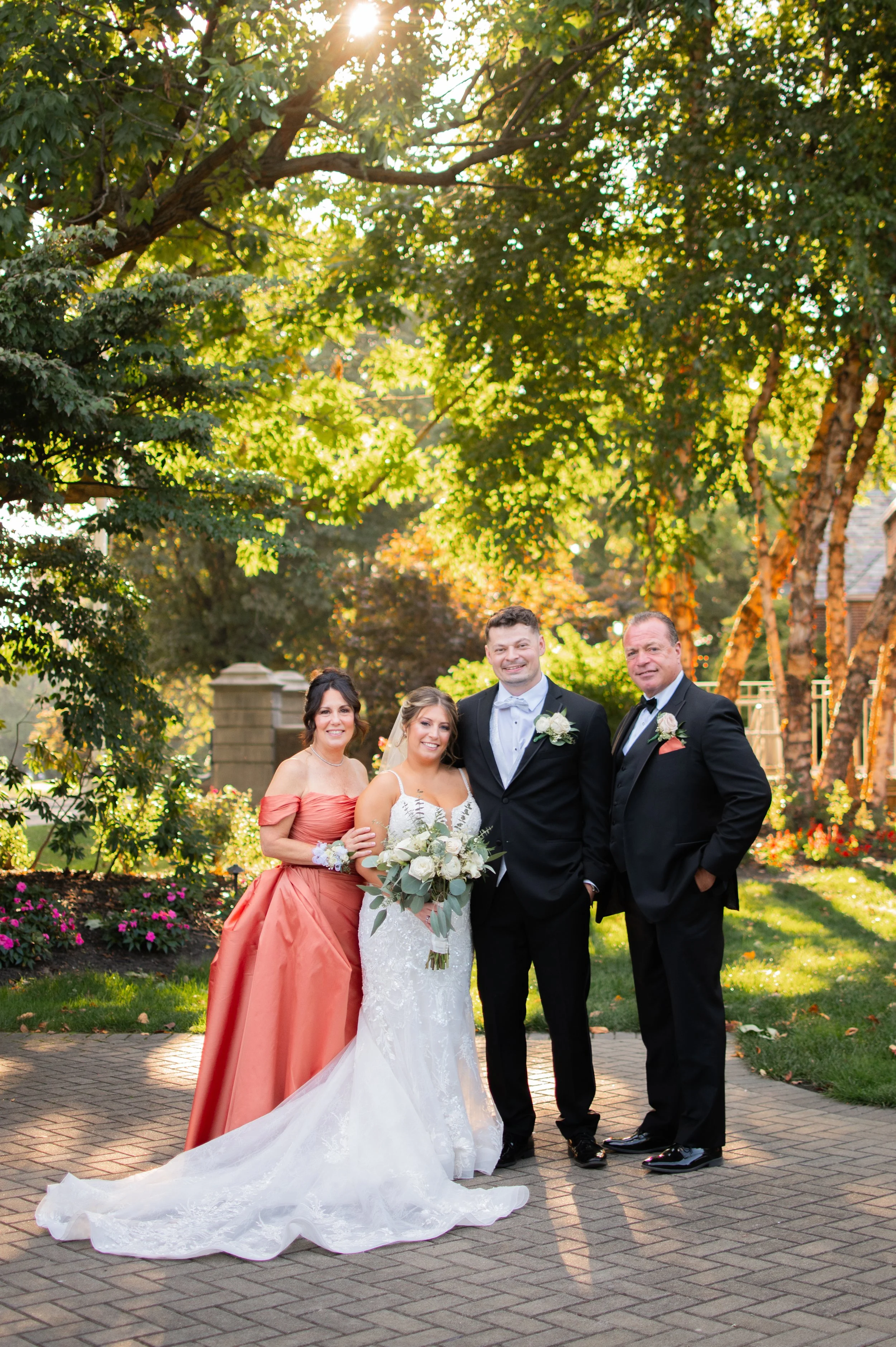 A group of five people, including a bride in a wedding dress with a train and bouquet, and four other adults dressed in formal attire, posing outdoors in a garden with trees and flowers on a sunny day.