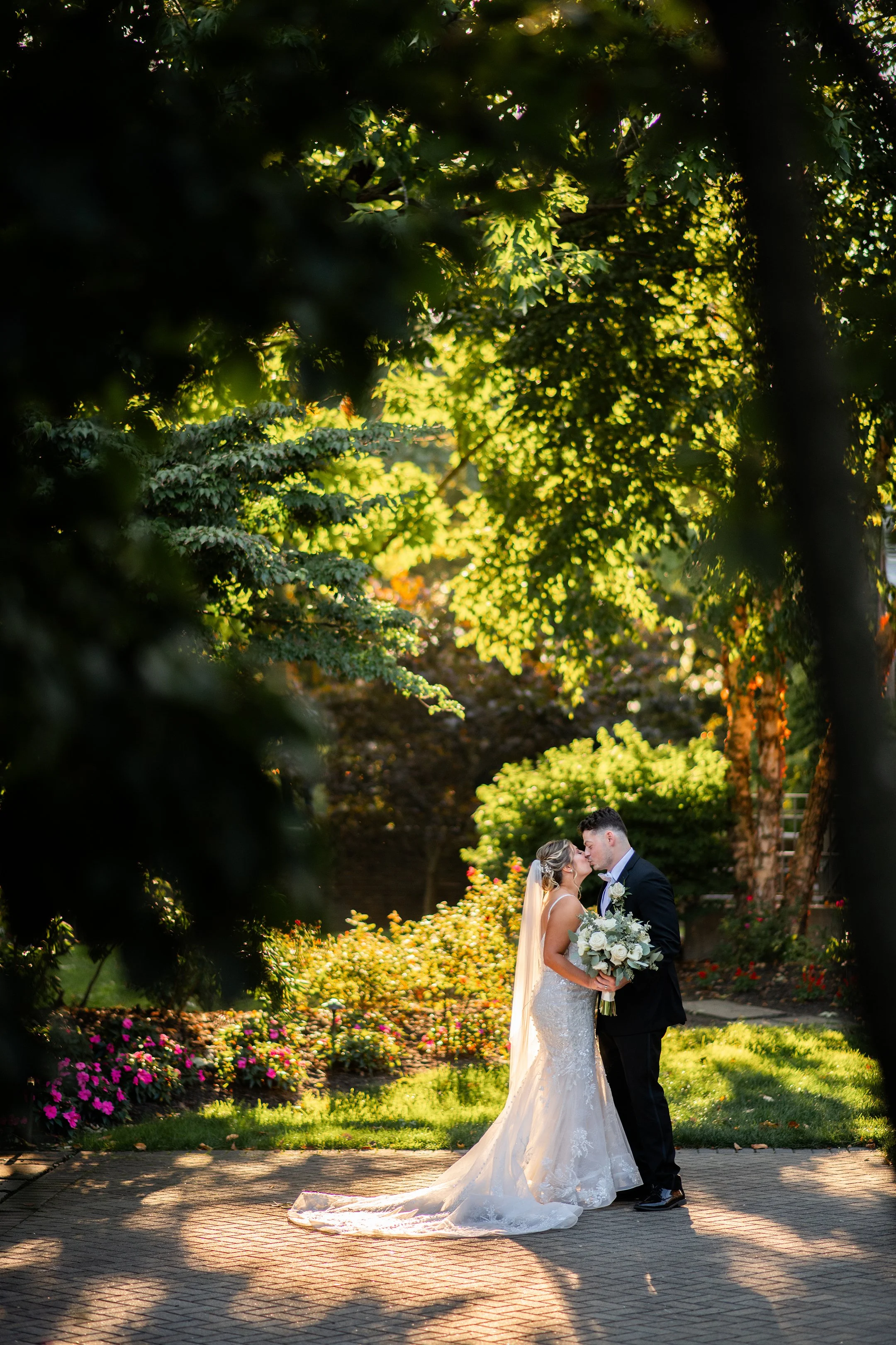 A bride and groom kissing outdoors in a garden with lush greenery and colorful flowers, surrounded by sunlight filtering through trees.