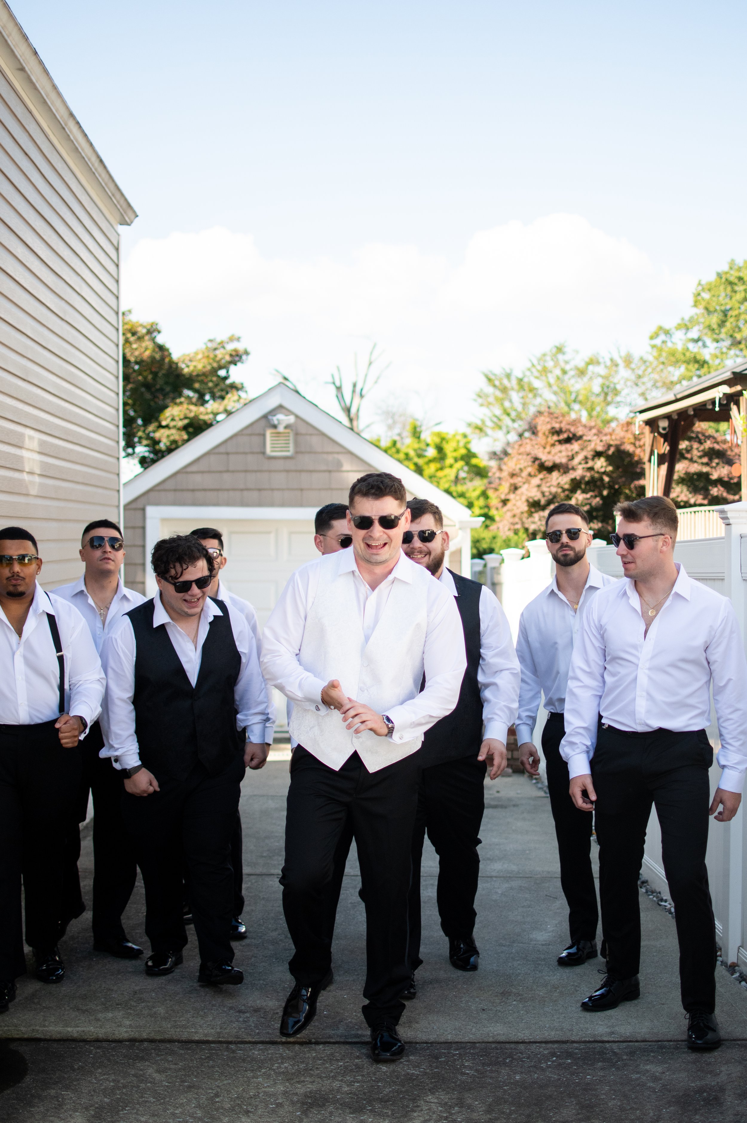 A group of men walking on a sidewalk outdoors, dressed in formal attire with white shirts and black pants, some wearing sunglasses, smiling and talking.