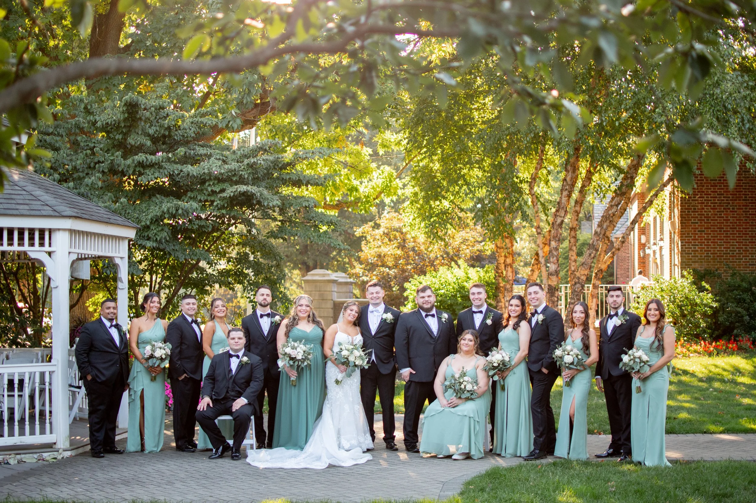 A group of people dressed in formal attire, including men in tuxedos and women in matching green dresses, posing outdoors with trees and a brick building in the background. It appears to be a wedding or formal event.