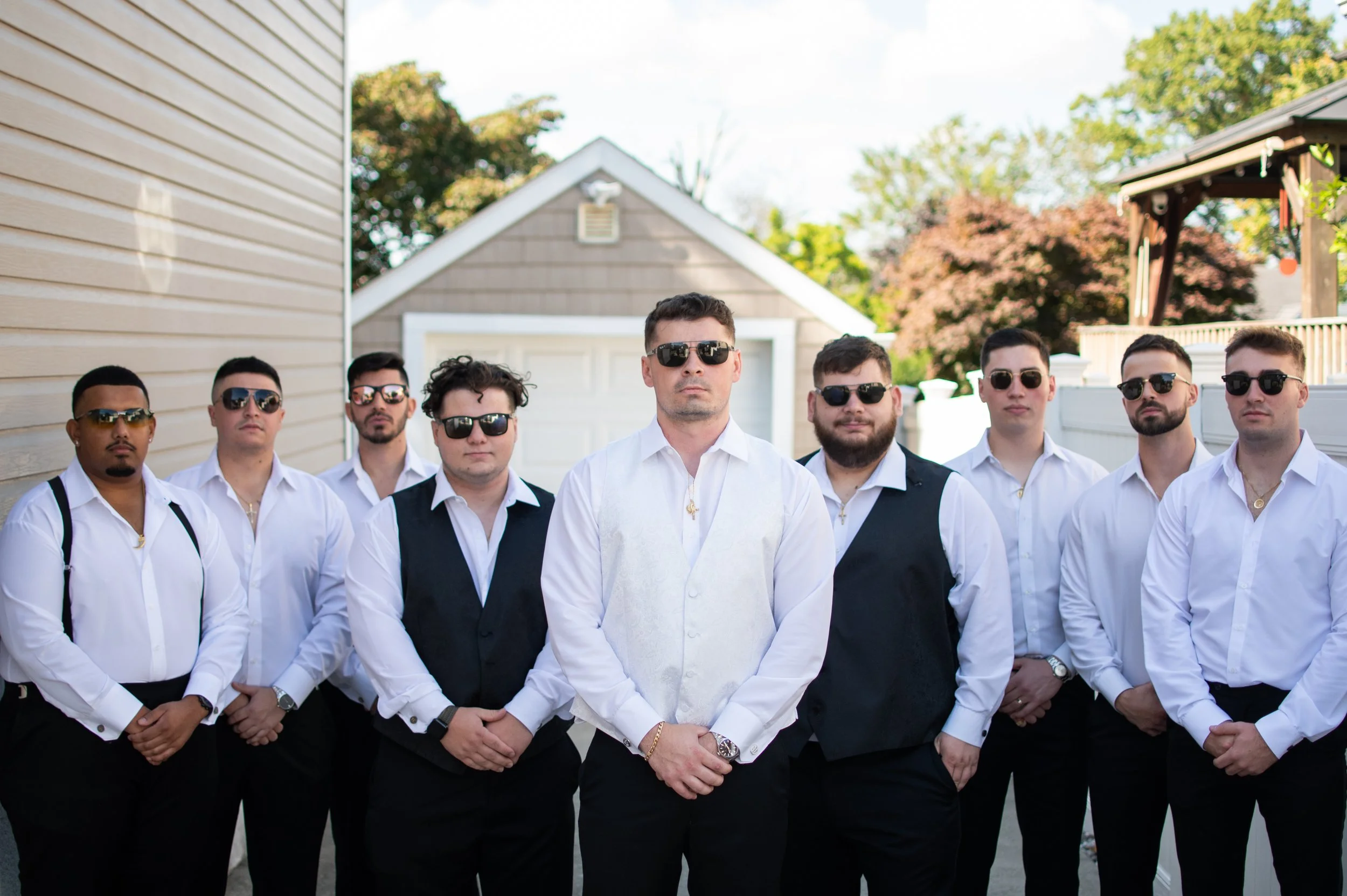 Group of men dressed in white shirts and black pants, wearing sunglasses, standing outdoors on a sunny day, with houses and trees in the background.