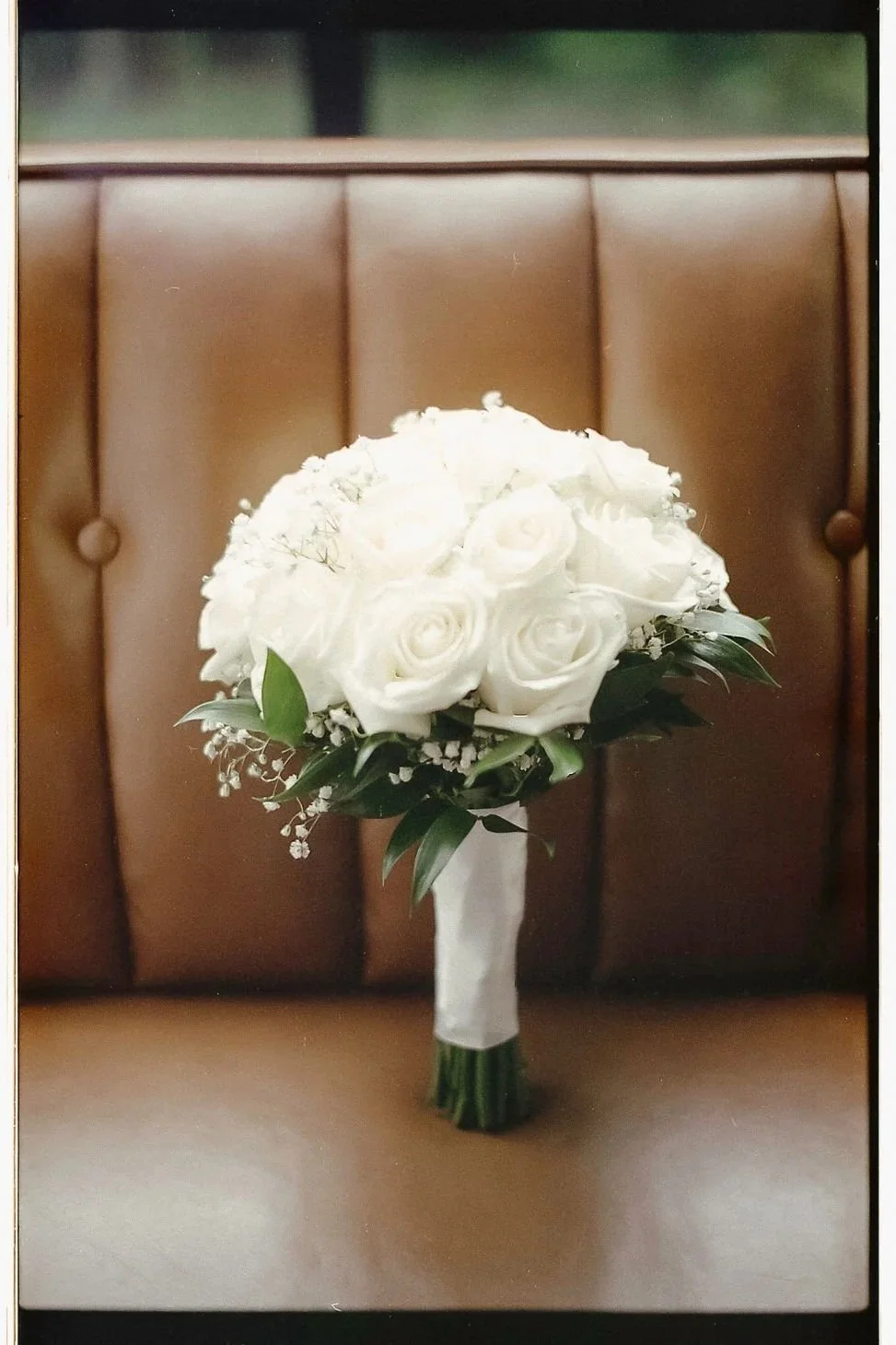 A wedding bouquet with white roses, baby's breath, and greenery, wrapped in white ribbon, placed on a wooden surface against a padded brown background.