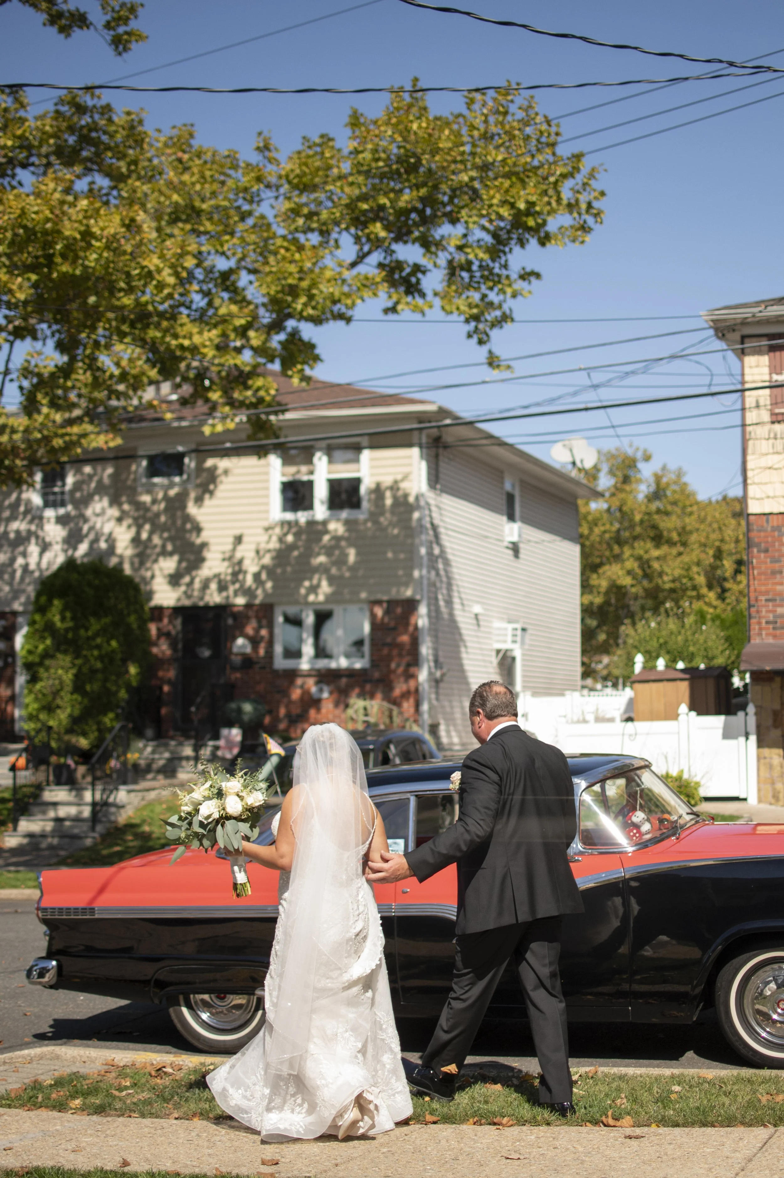 A bride in a white wedding dress holding a bouquet of white flowers and greenery, standing outside next to a vintage red and black car, with a man in a black suit holding her hand on a sunny day with a blue sky, trees, and residential houses in the b