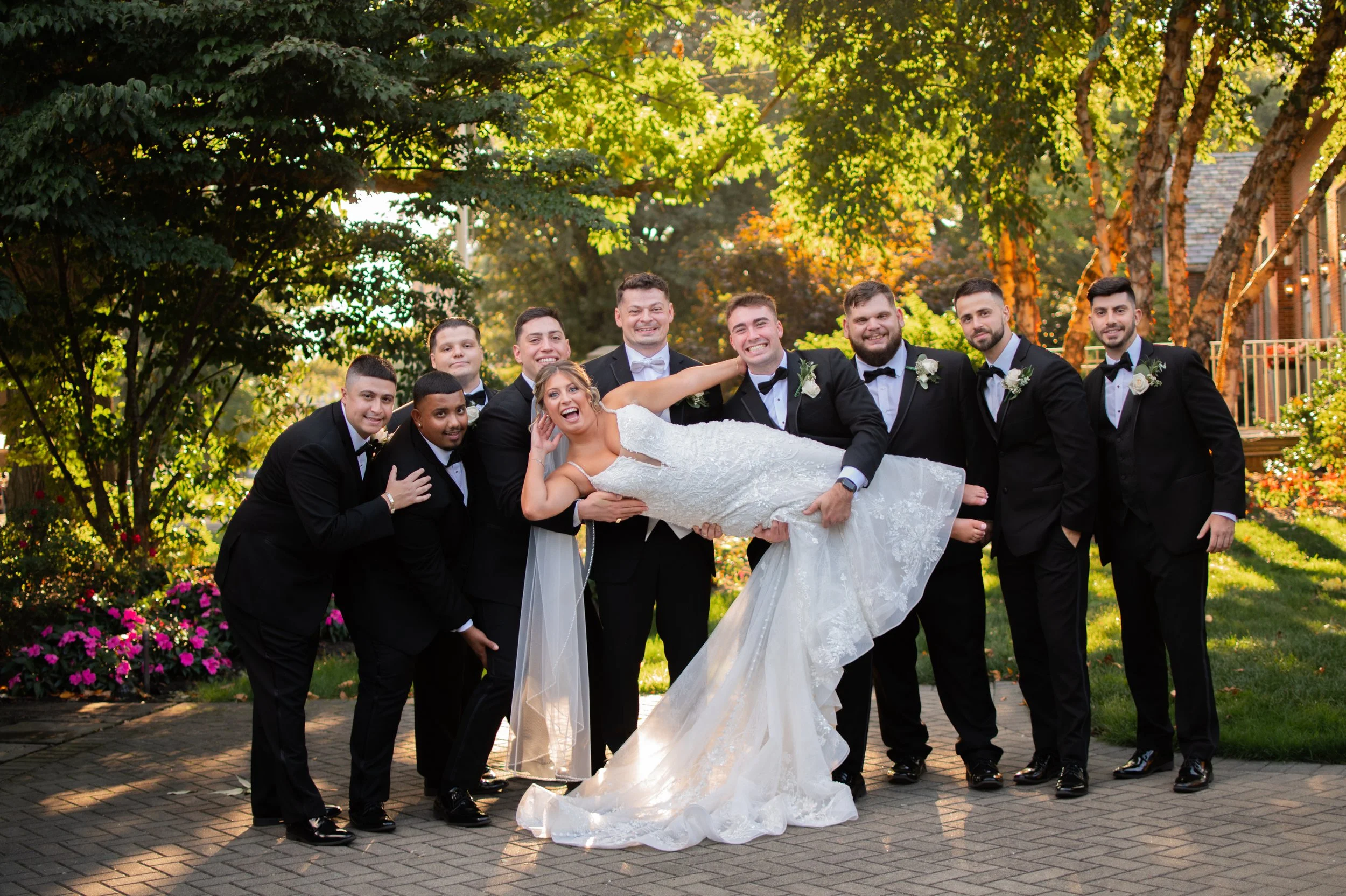 A bride in a white wedding gown lying in the arms of a groom in a black tuxedo, surrounded by four groomsmen in black tuxedos, posing outdoors beneath trees with sunlight. The group is smiling and celebrating.