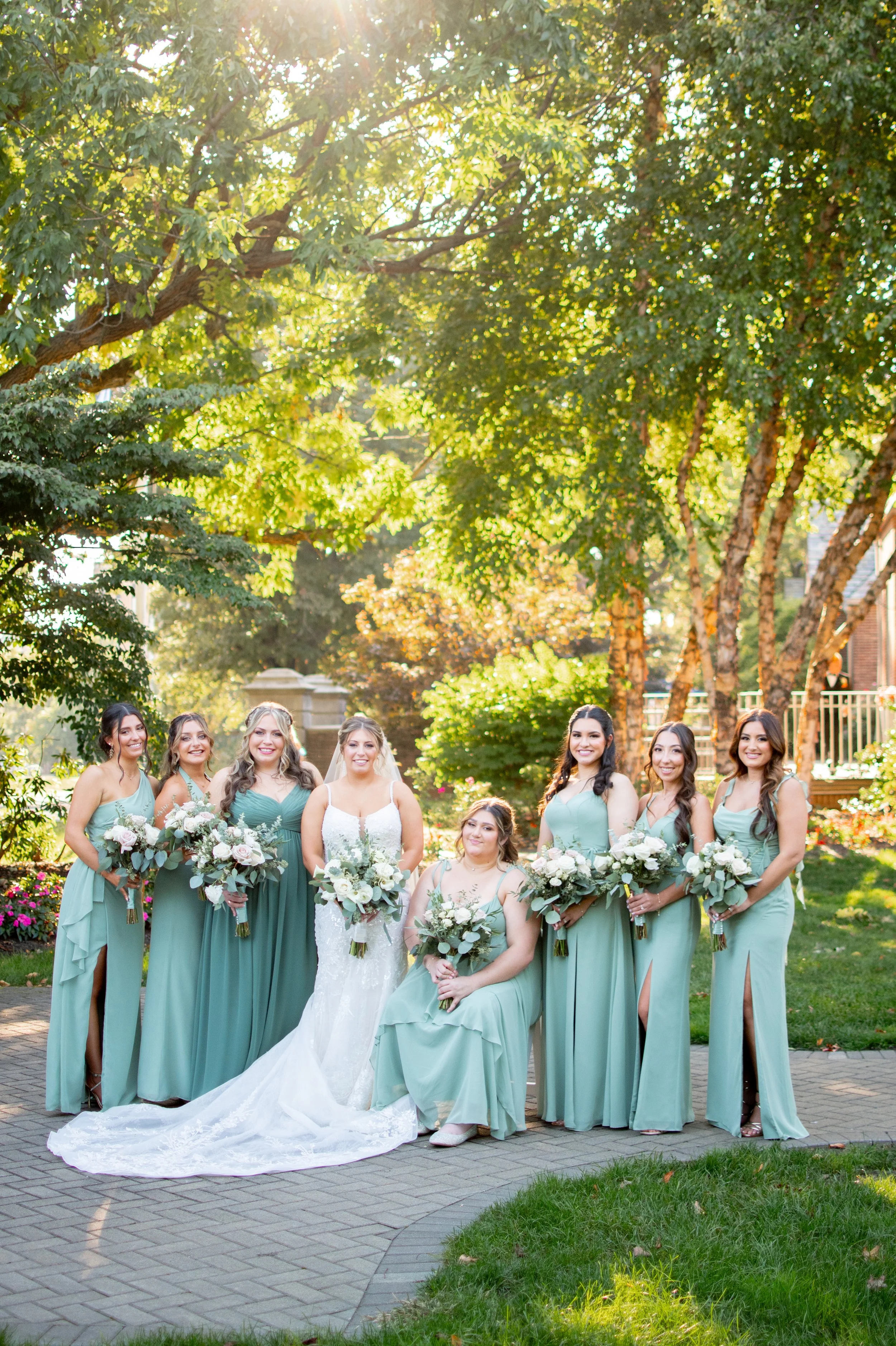 A group of women in mint green dresses and a bride in a white wedding gown standing outdoors under trees, holding bouquets of flowers during a wedding celebration.