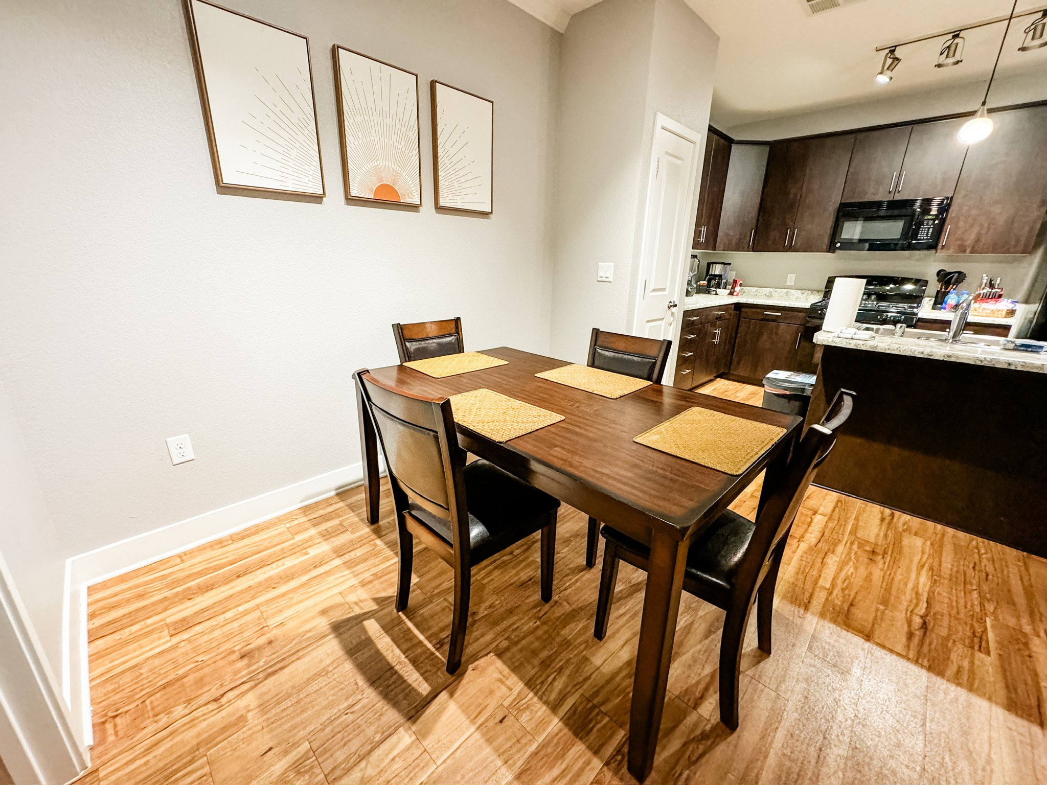Dining area and kitchen with a wooden dining table and four chairs, four placemats, and kitchen cabinets with appliances, counter, and a roll of paper towels.