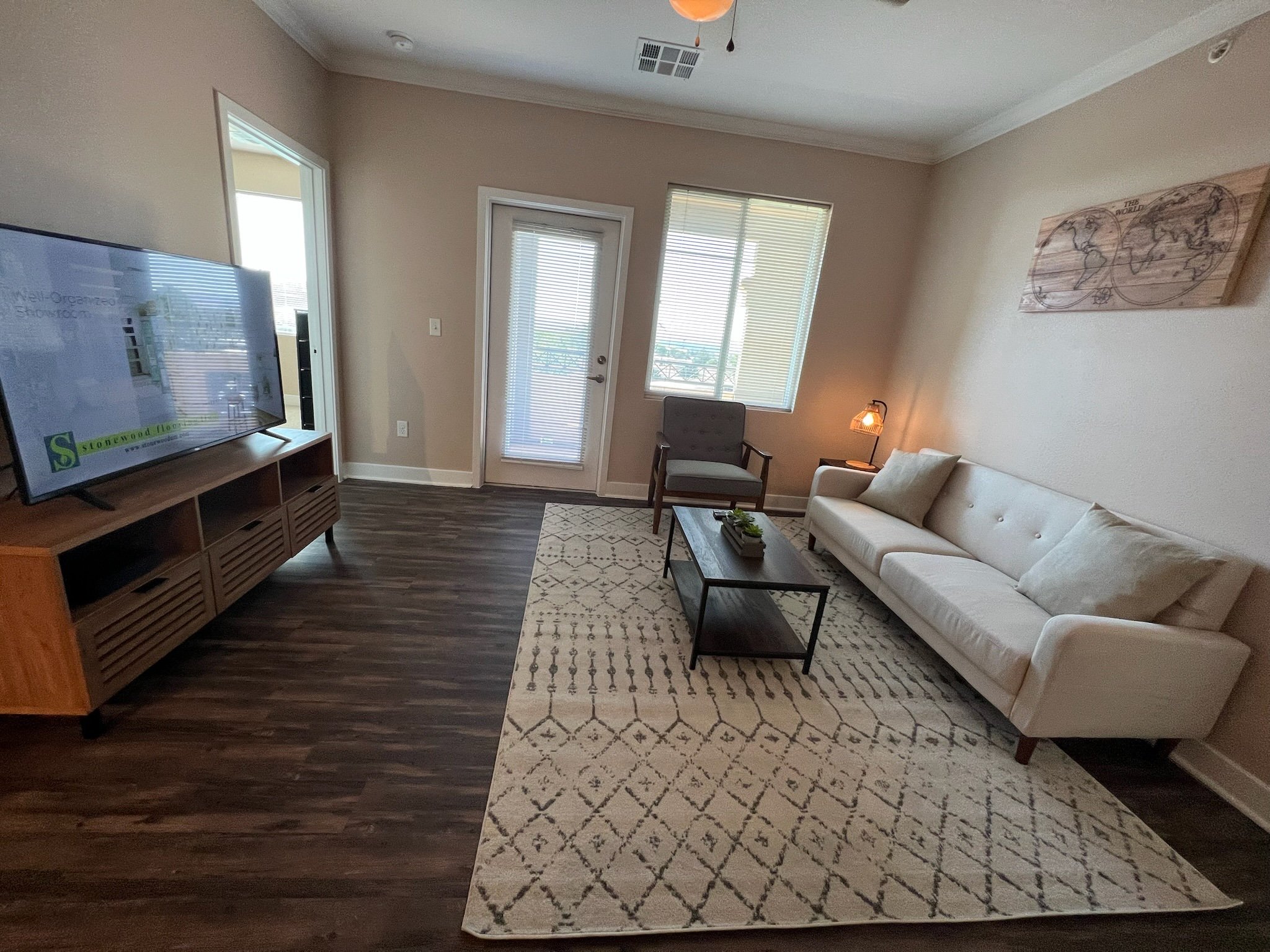 Living room with white sofa, gray armchair, coffee table, TV on wooden stand, and patterned rug, with door and windows allowing natural light.