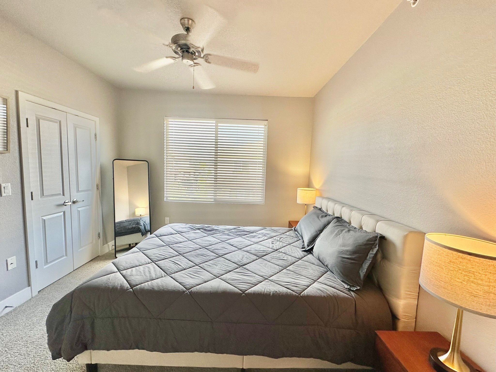 A bedroom with a bed, pillows, nightstand, lamps, window with blinds, closet with double doors, a ceiling fan, and a mirror.