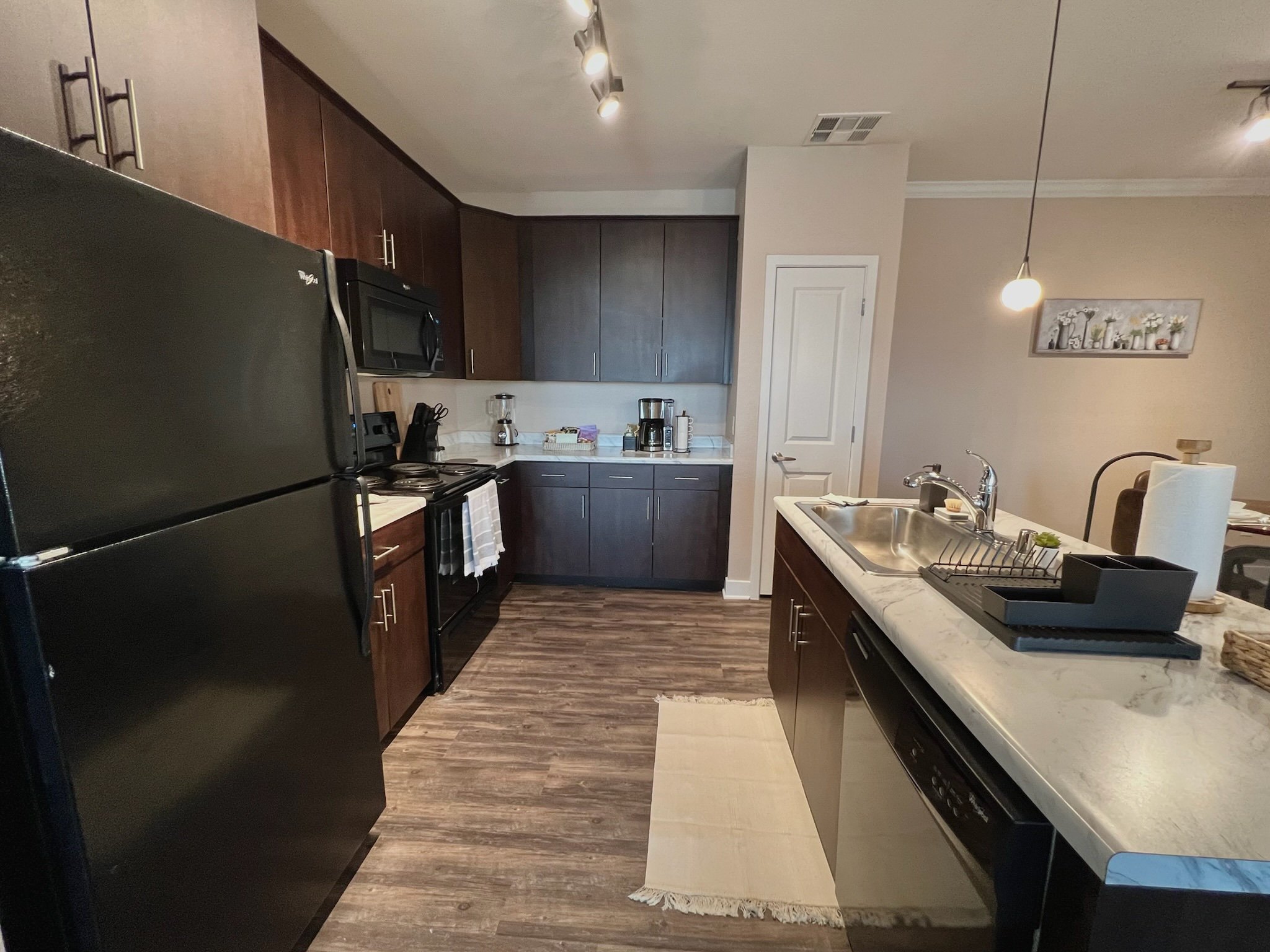 Kitchen with dark brown cabinets, a black refrigerator, a stove, microwave, and coffee maker. There is a kitchen island with a sink, a paper towel roll, and a dish rack. The wood flooring runs throughout the space, and there's a beige wall with a fra