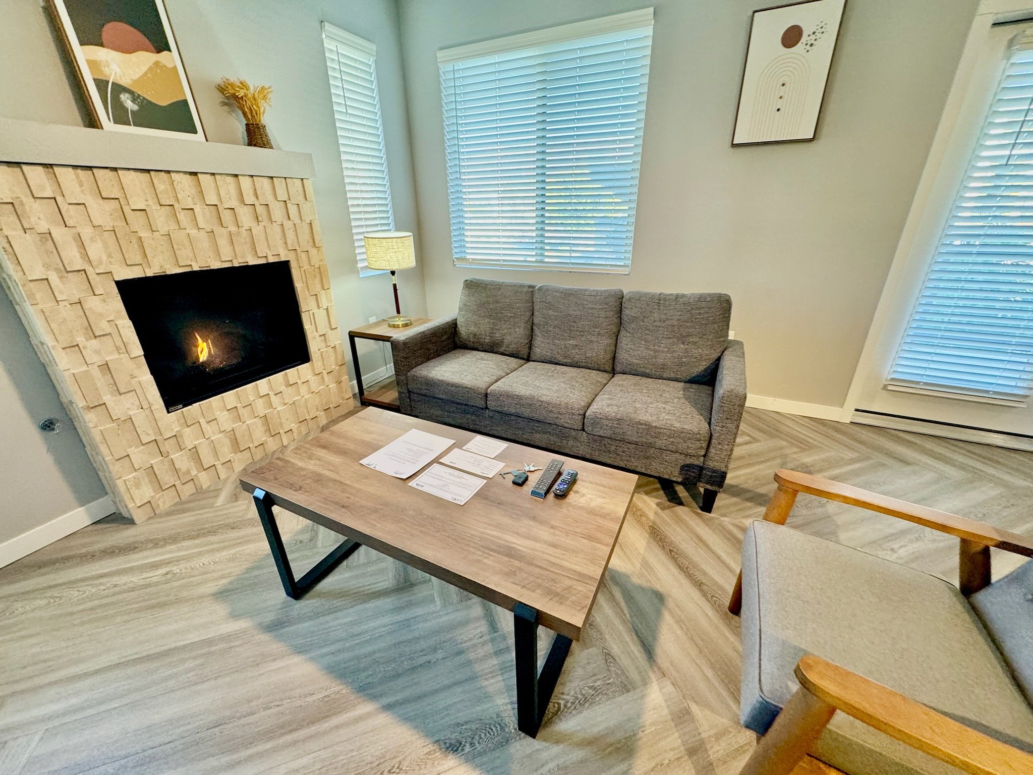 Living room with gray sofa, wooden coffee table with papers, remote controls, fireplace with beige wooden surround, windows with blinds, artwork on walls, small side table with lamp, beige armchair with wooden arms.