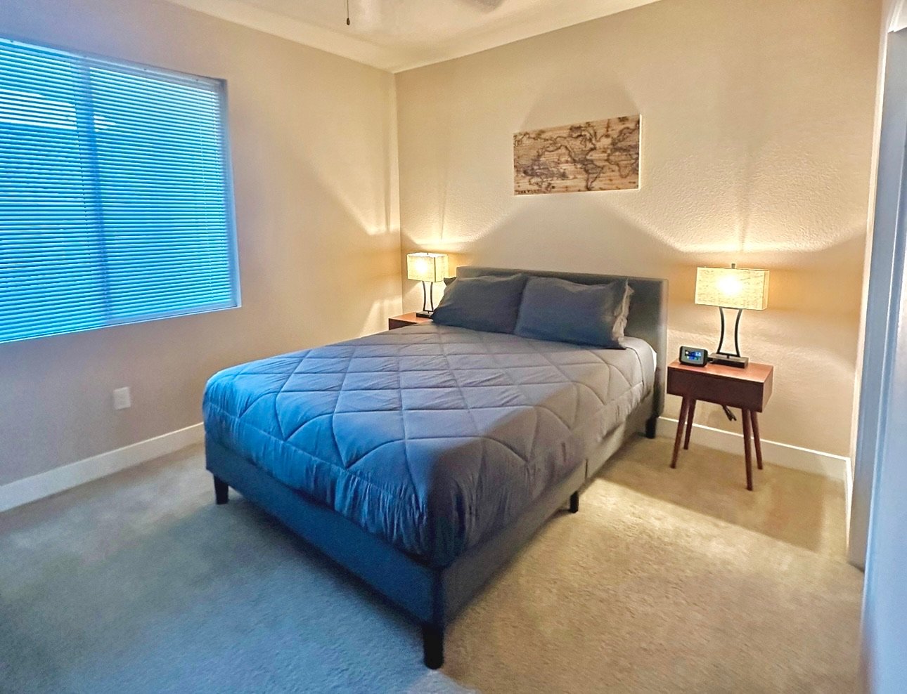 Bedroom with a king-sized bed, dark headboard, grey bedding and pillows, two wooden nightstands with lamps, a digital alarm clock, a world map art piece on the wall, beige walls, carpeted floor, and window with closed blinds.