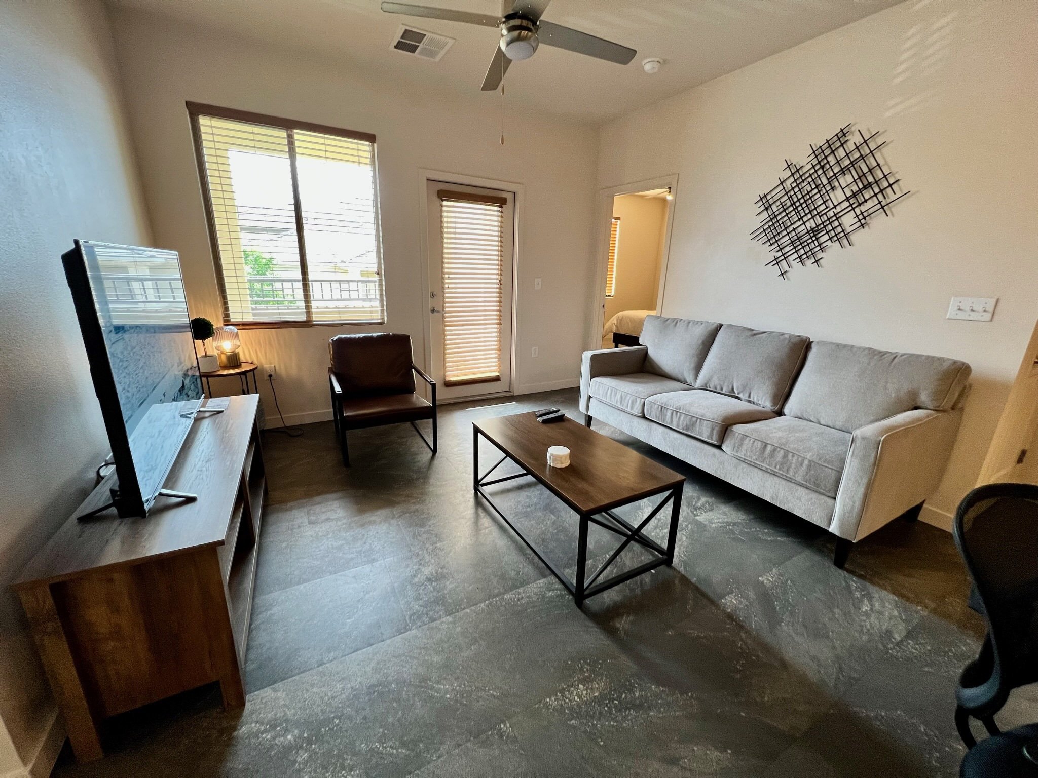 Living room with a gray sofa, wooden coffee table, television on a wooden stand, brown armchair, wall decor, window, door with blinds, ceiling fan, and a bedroom visible through an open door.