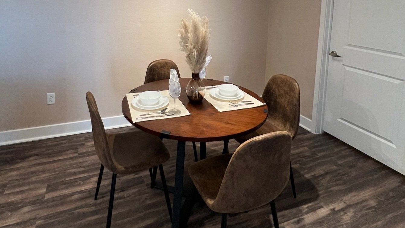 A round wooden dining table set for four with white plates, bowls, and silverware, decorated with a brown vase containing dried pampas grass, in a room with dark wood flooring and beige walls.