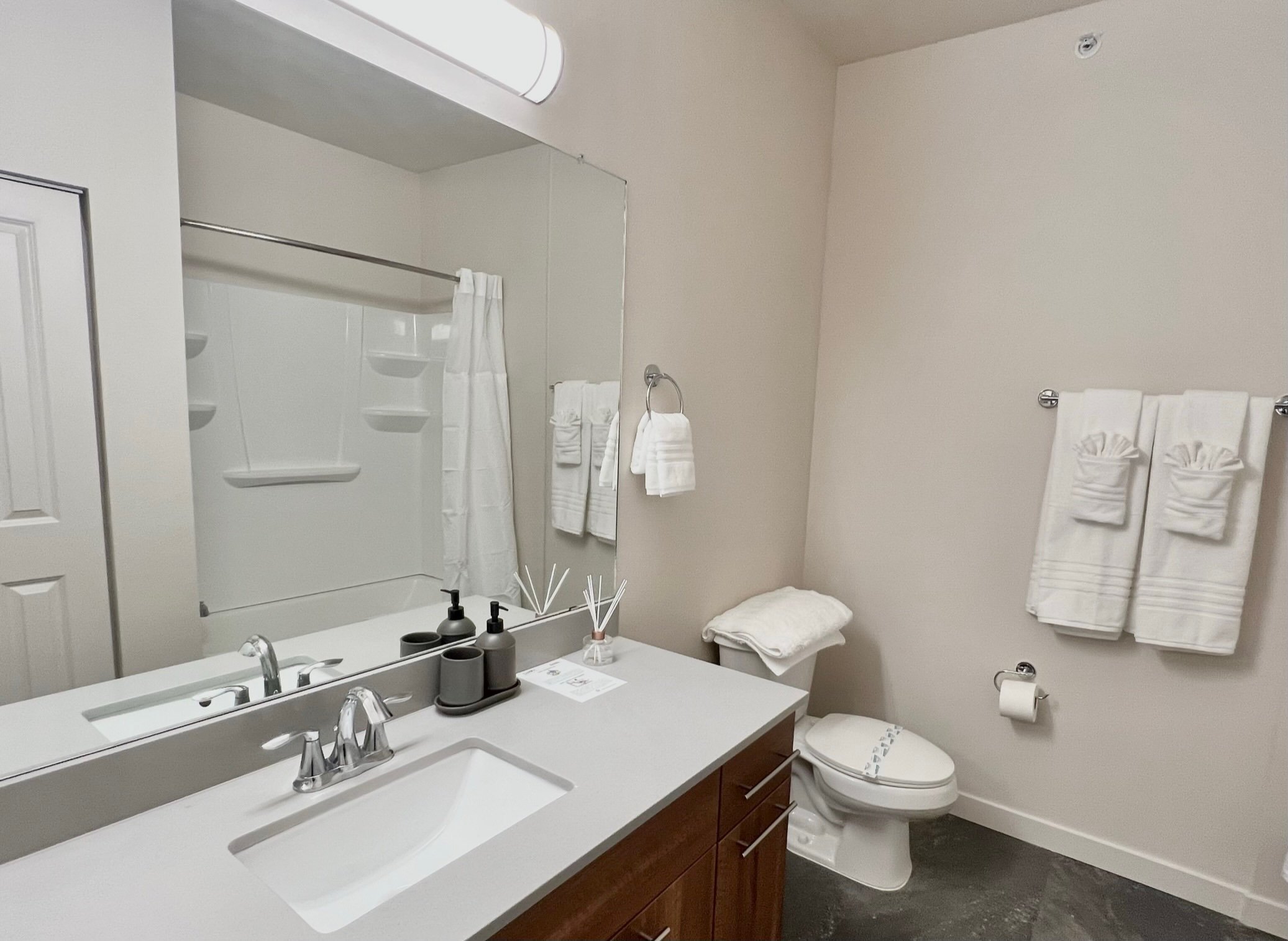 A clean bathroom with a white countertop, a sink with a silver faucet, soap dispensers, a decorative reed diffuser, a large mirror, and a beige wall. There are towel racks with neatly folded white towels, a toilet with a lid, and a shower area with a
