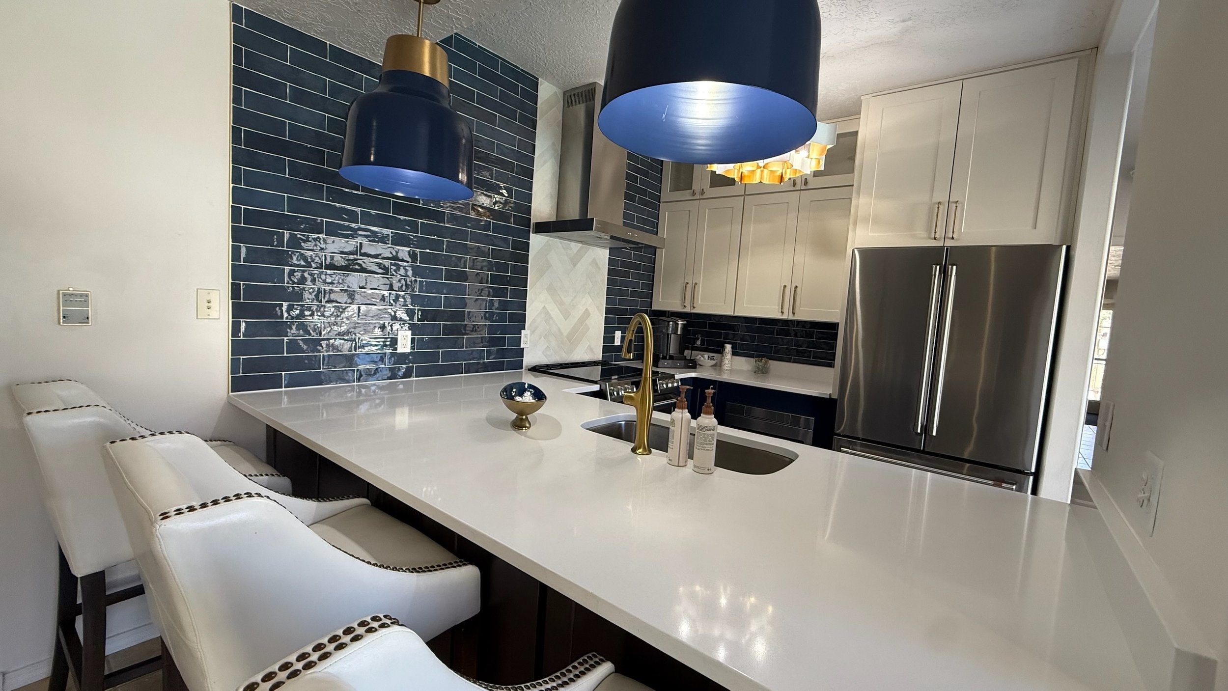 Modern kitchen with white countertops, navy blue tile backsplash, white cabinets, stainless steel refrigerator, and black pendant lights.