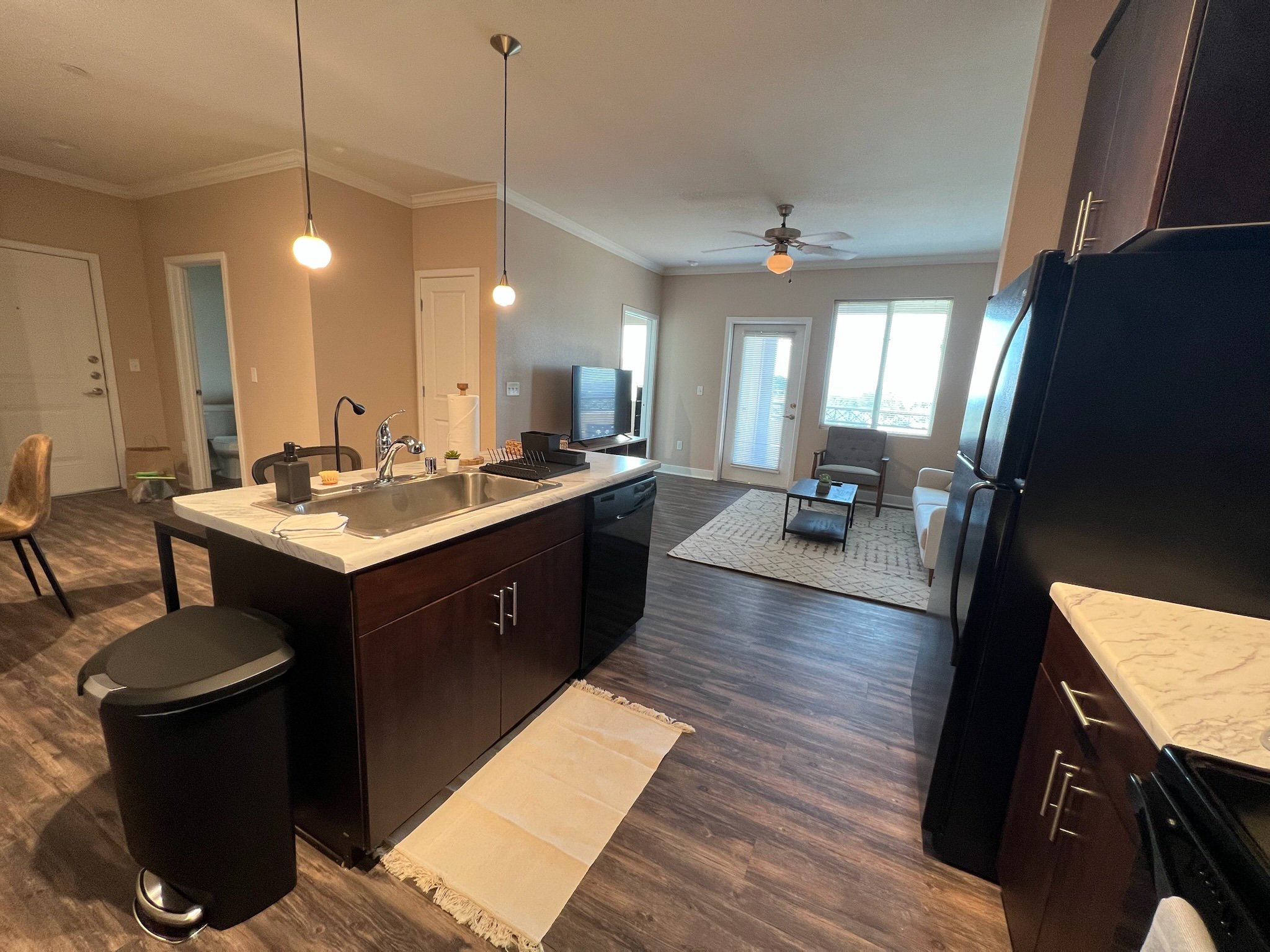 Open-concept living space with kitchen island, dining area, and living room with sofa and armchair, illuminated by natural light from large windows and balcony door.