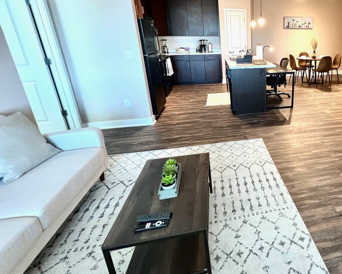 Living room with beige couch, dark wood coffee table with potted succulents, patterned rug, and TV remote. Open kitchen with dark cabinetry, refrigerator, and dining area with four chairs in the background.